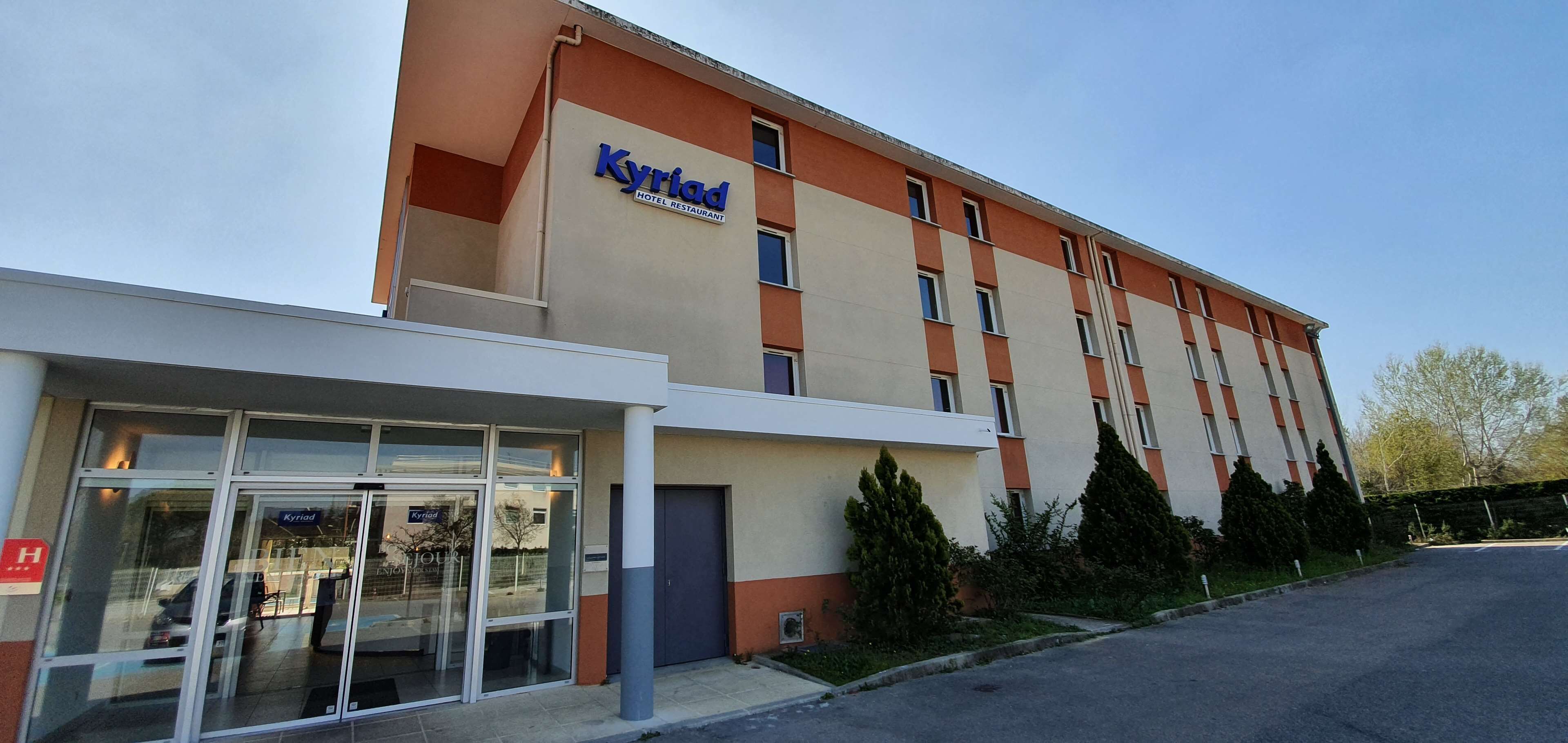 Um hotel com fachada moderna, entrada de vidro e uma placa com a inscrição "Kyriad Hotel Restaurant".