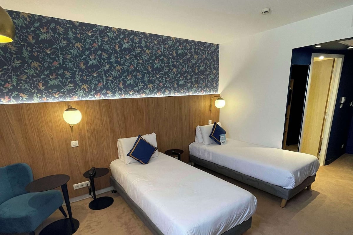 A hotel room with two single beds, a small desk, a chair, and a decorative wall.