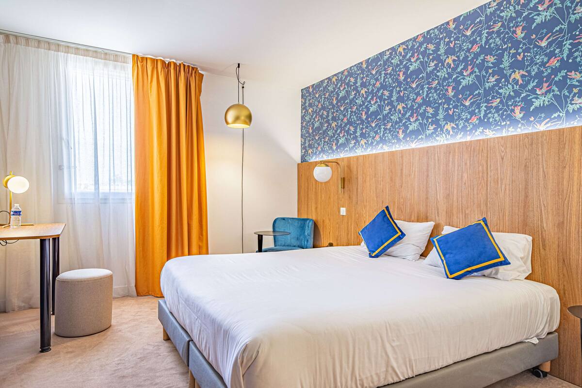 A hotel room with a large bed, a desk with a lamp, a chair, and a round ottoman.