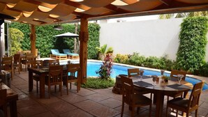 Un área de comedor al aire libre con mesas y sillas de madera, una piscina y abundante vegetación.
