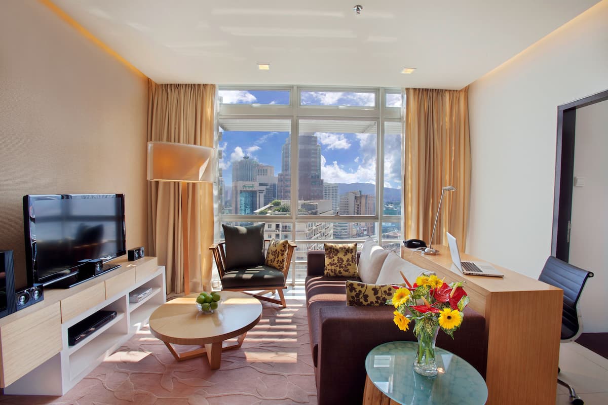 Ein modernes Hotelzimmer mit großem Fernseher, bequemen Sitzgelegenheiten, einem Schreibtisch mit Laptop und Blick auf die Stadtlandschaft.