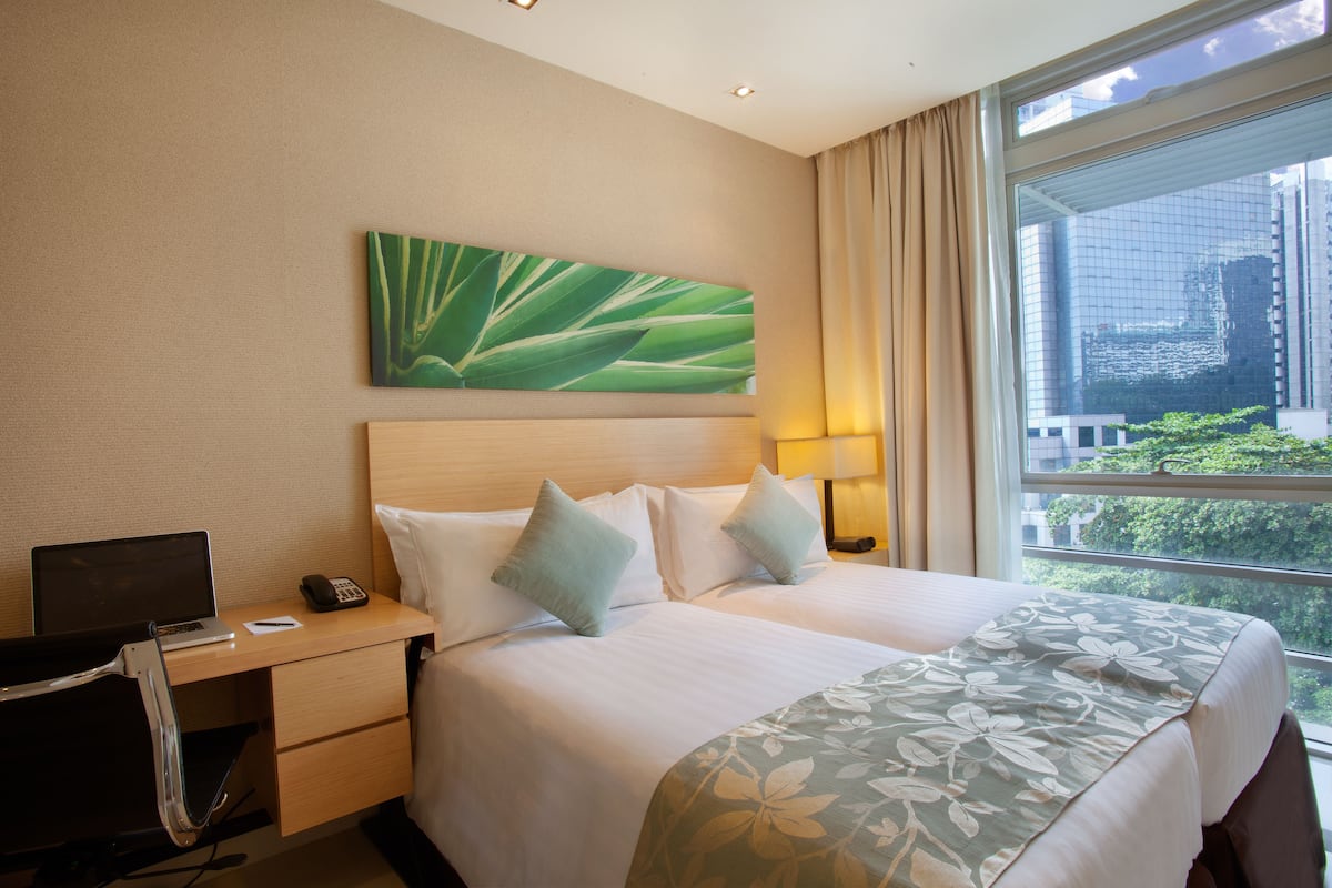 Ein Hotelzimmer mit einem Bett, einem Schreibtisch mit Laptop und Telefon, einem Bild an der Wand und einem Fenster mit Blick auf Gebäude und Grünflächen.