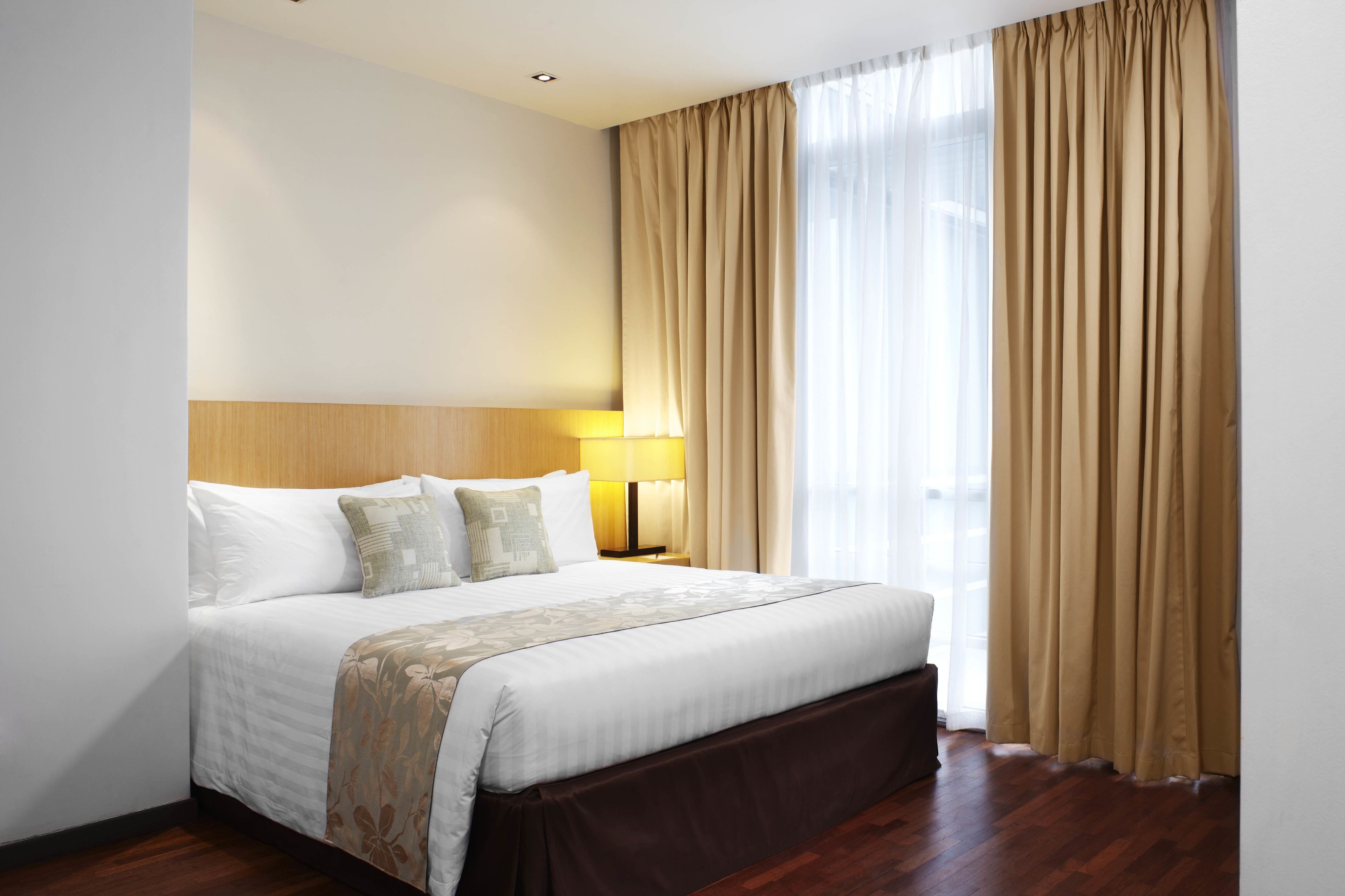 Habitació d'hotel amb un llit gran, un panell de fusta al capçal, una tauleta de nit amb una làmpada i vista a l'exterior a través de cortines translúcides.