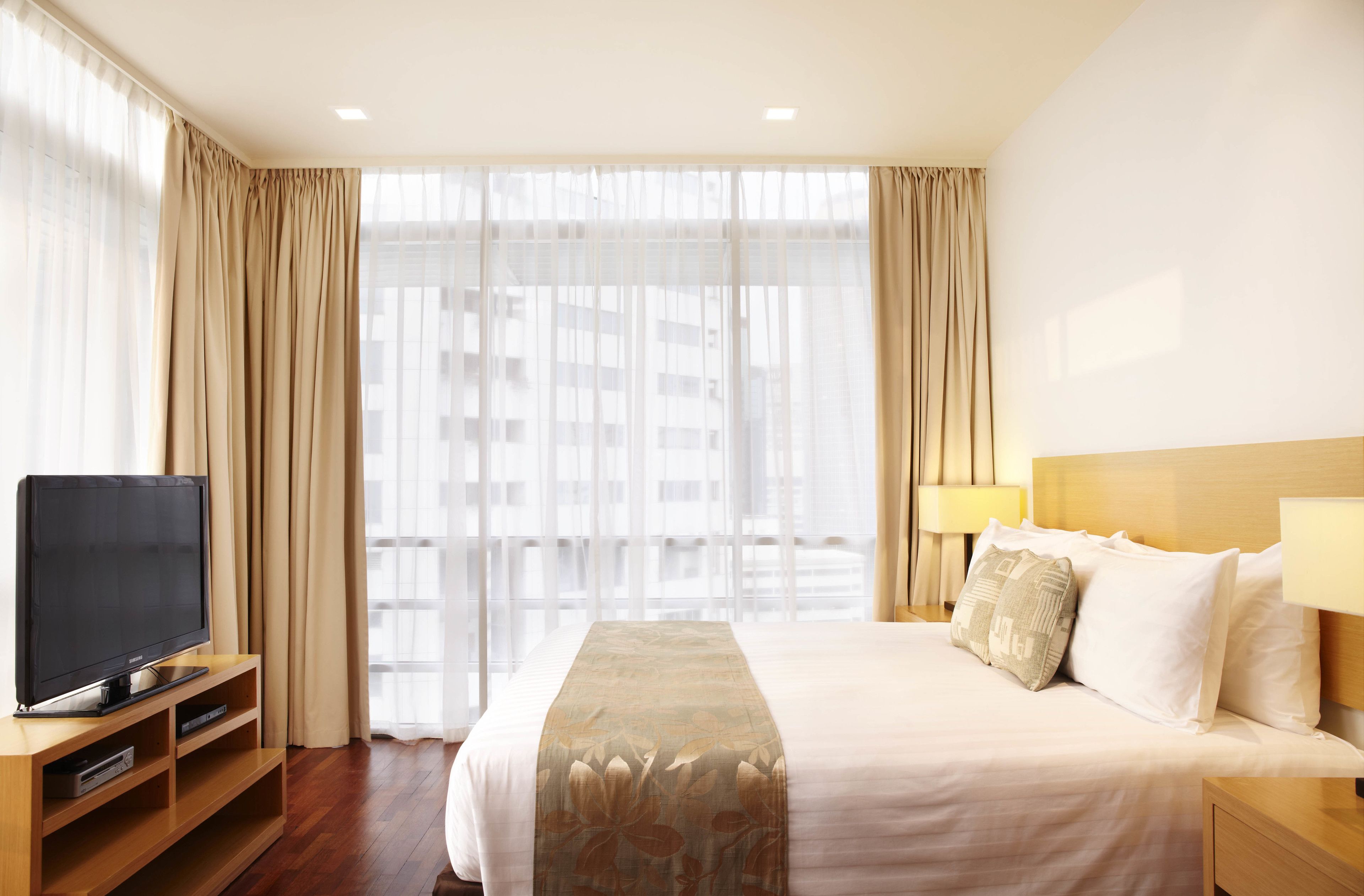 A hotel room with a bed, a TV on a wooden stand, and a window with sheer curtains.