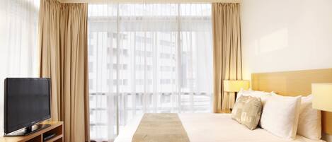 A hotel room with a bed, a TV on a wooden stand, and a window with sheer curtains.