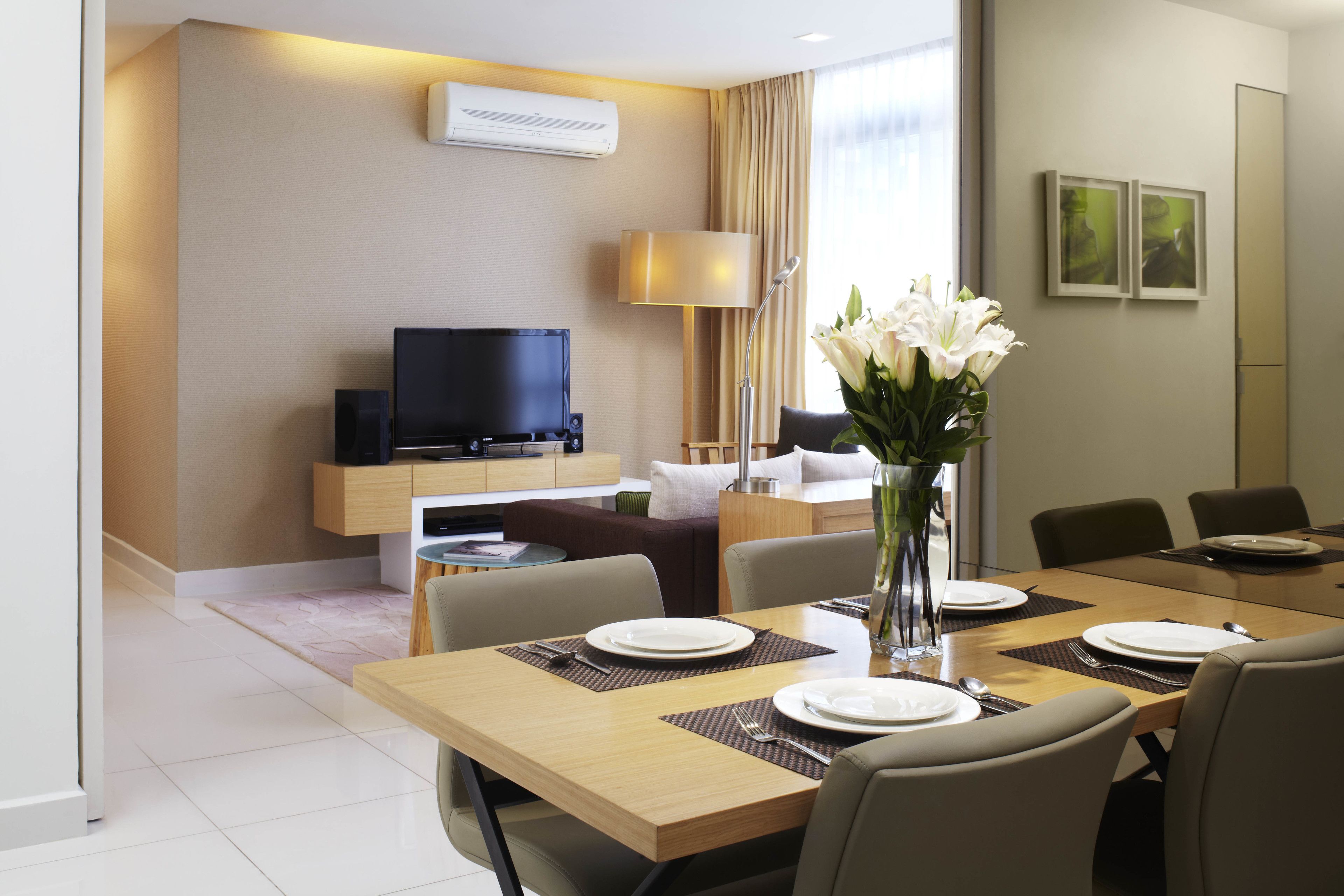 A hotel dining area with a wooden table set for four, a television in the background, and a vase with white flowers on the table.
