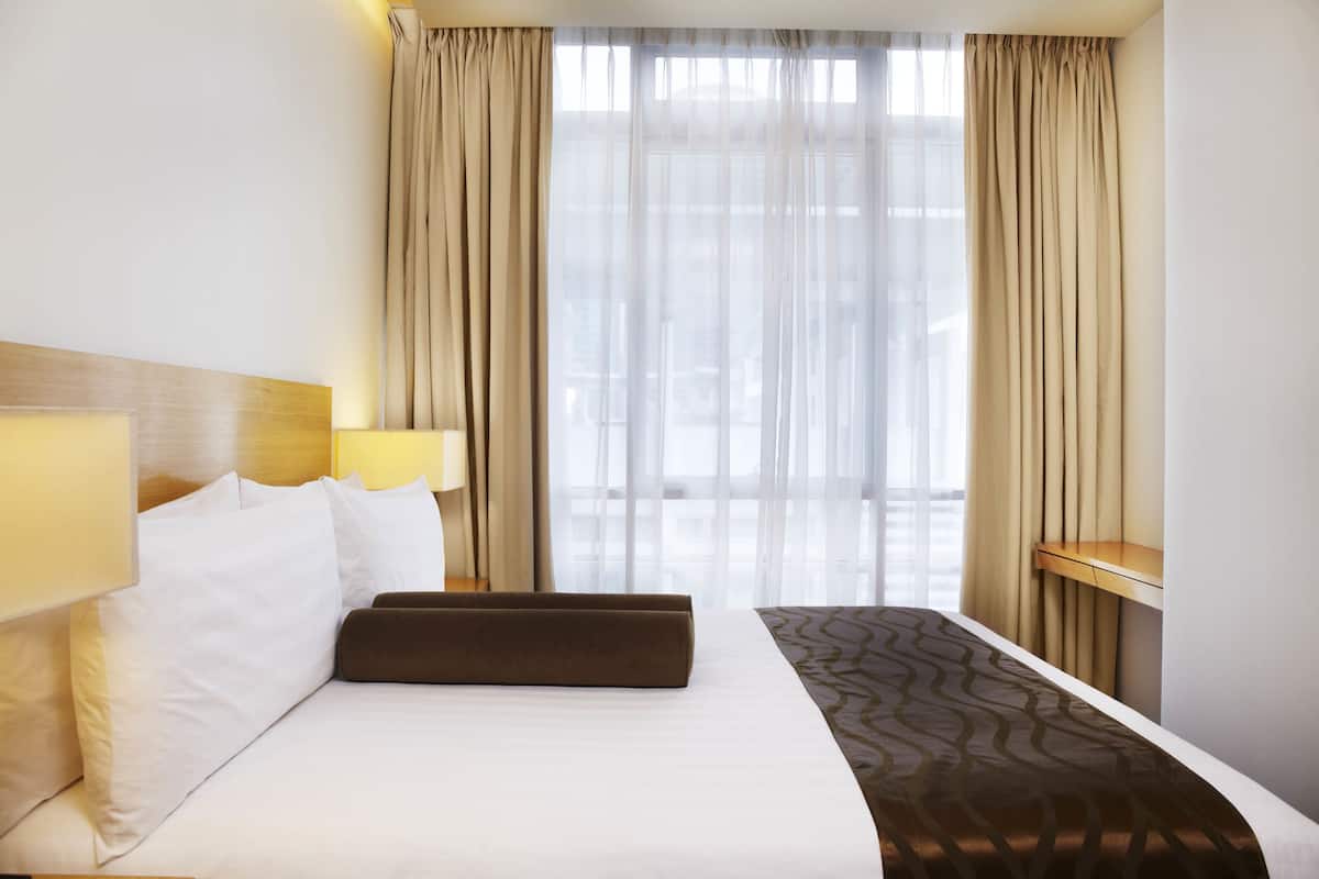 Ein Hotelzimmer mit einem großen Bett, zwei Nachttischlampen, einem Holz-Kopfteil, einem Fenster mit durchsichtigen Vorhängen und Blick auf die Stadt.