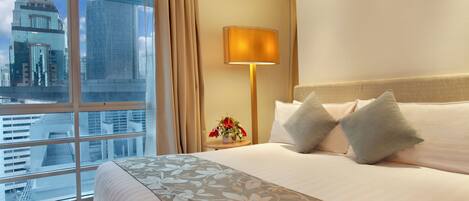 A hotel room with a large bed, a bedside lamp, a small table with a vase of flowers, and a view of city buildings through a large window.