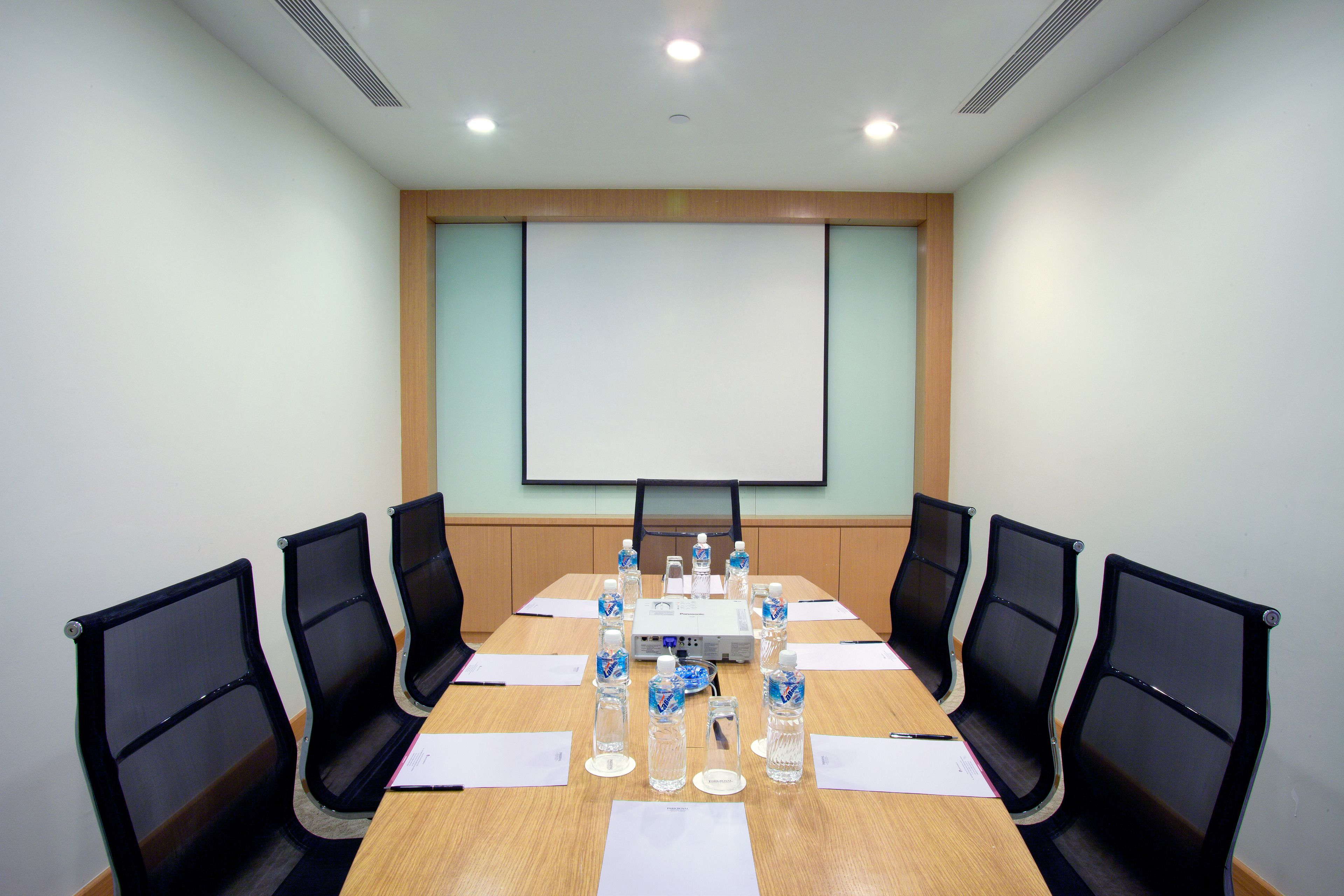 A conference room with a long table, black chairs, water bottles, and a projector screen.