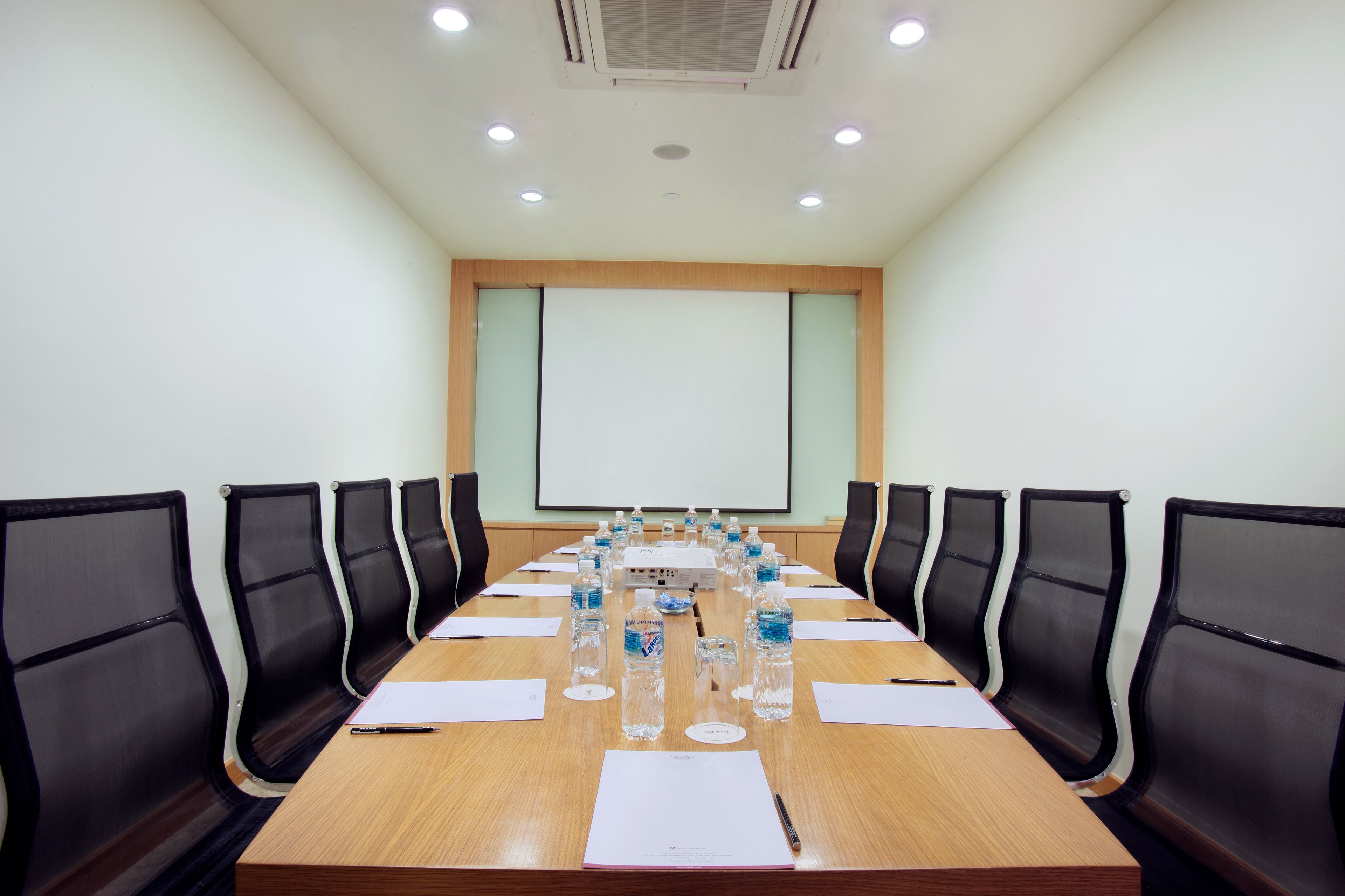 A conference room with a long table, black chairs, water bottles, and a projector screen.