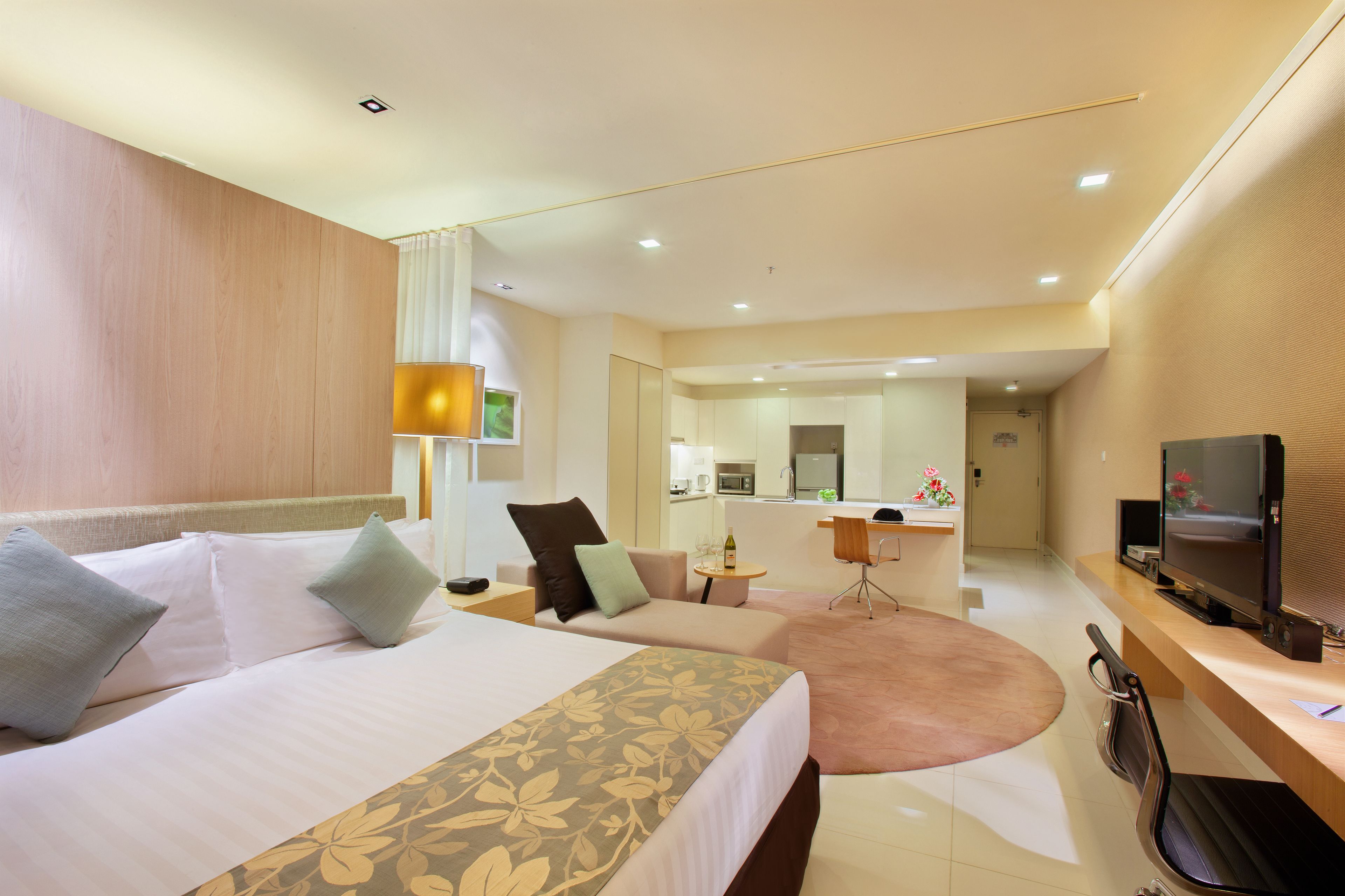 A hotel room with a bed, a desk, a TV, and a kitchenette in the background.