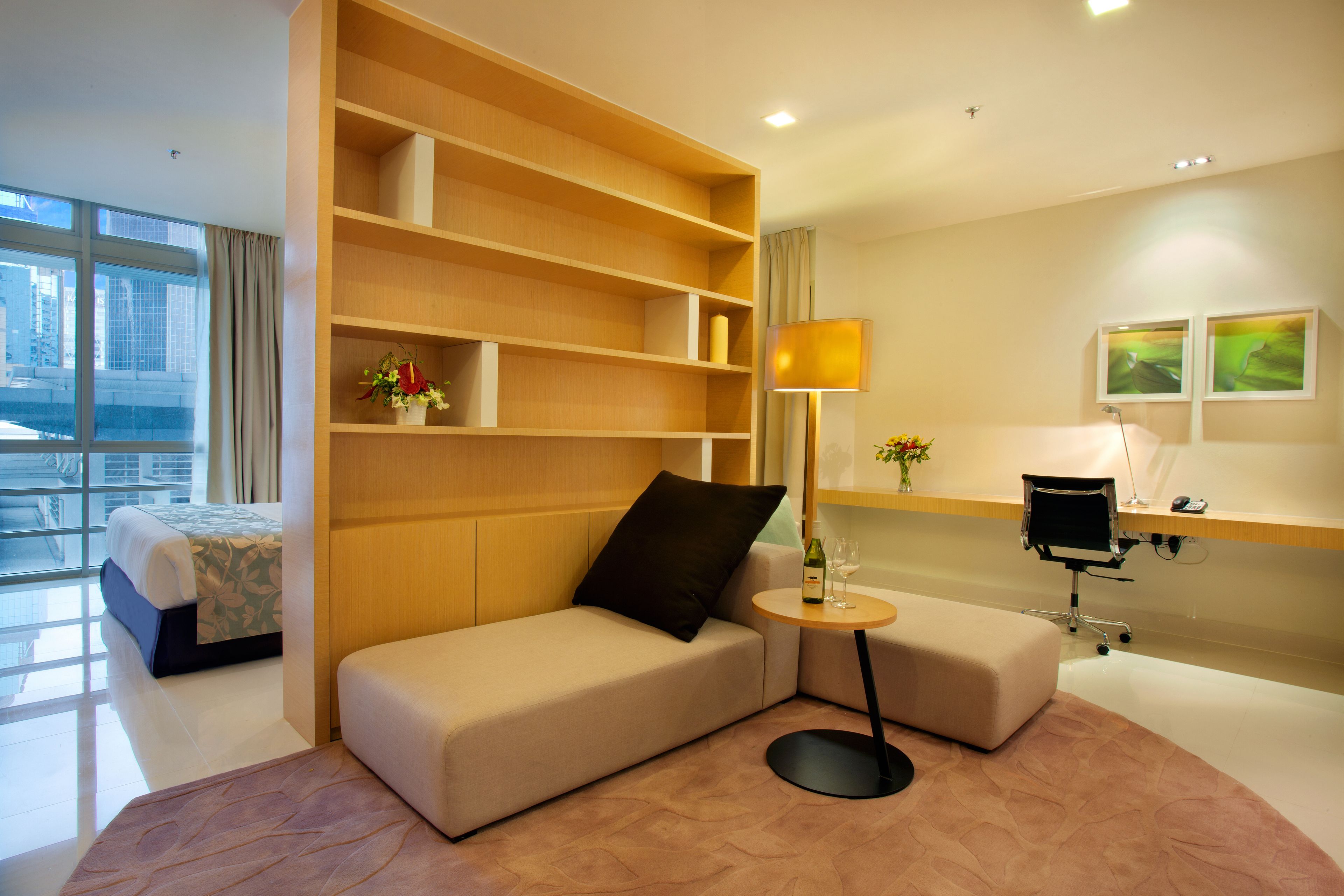 A modern hotel room with a sofa, a small round table with a lamp, a desk with a chair, and a large window offering a city view.