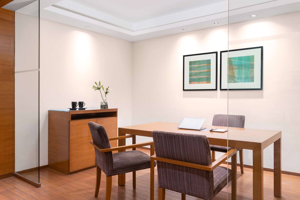 A conference room with a wooden table, four chairs, a glass partition, and two framed abstract paintings on the wall.