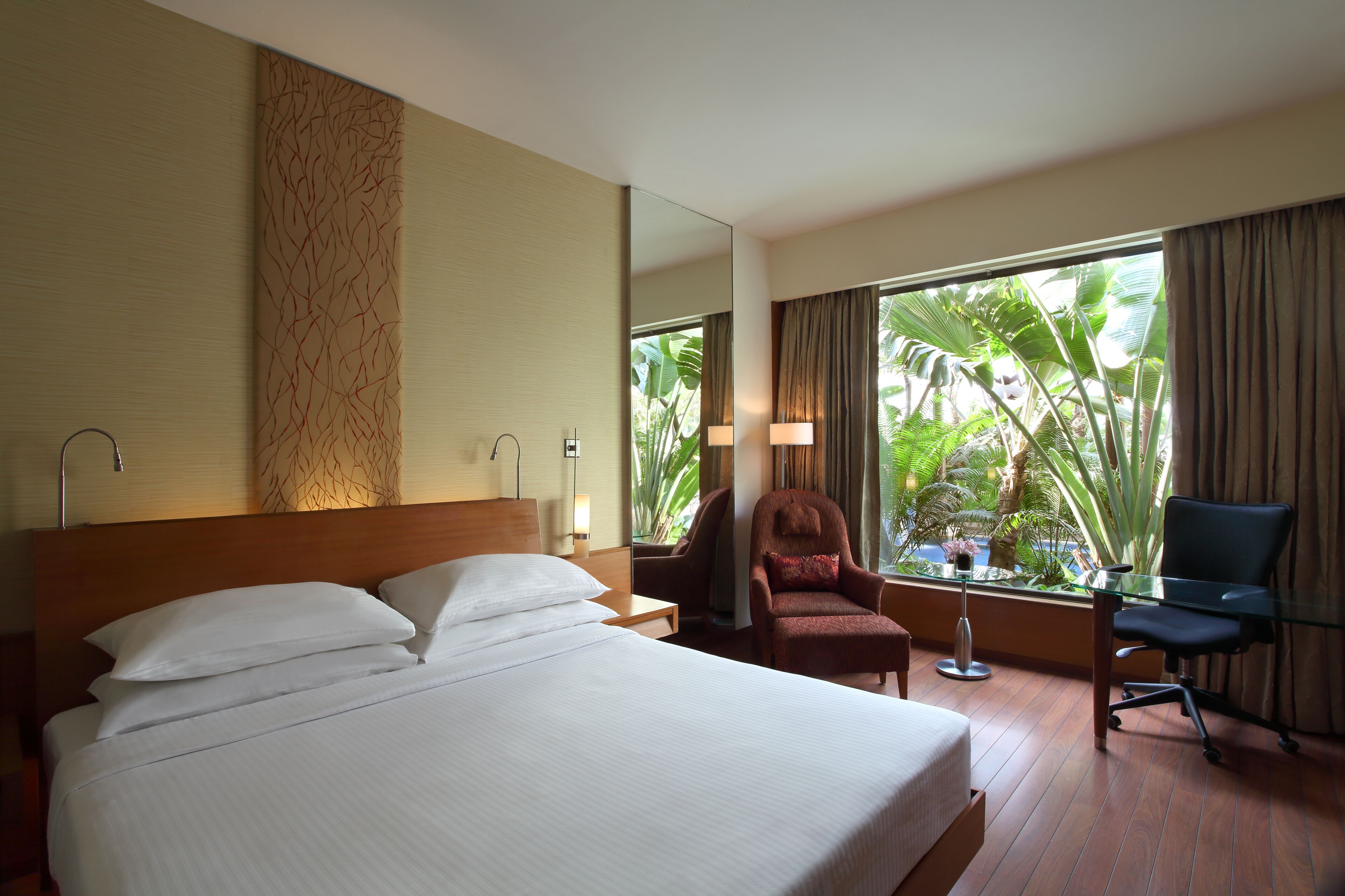 Une chambre d’hôtel avec un grand lit, un bureau avec une chaise, une petite table et une fenêtre donnant sur une végétation tropicale.