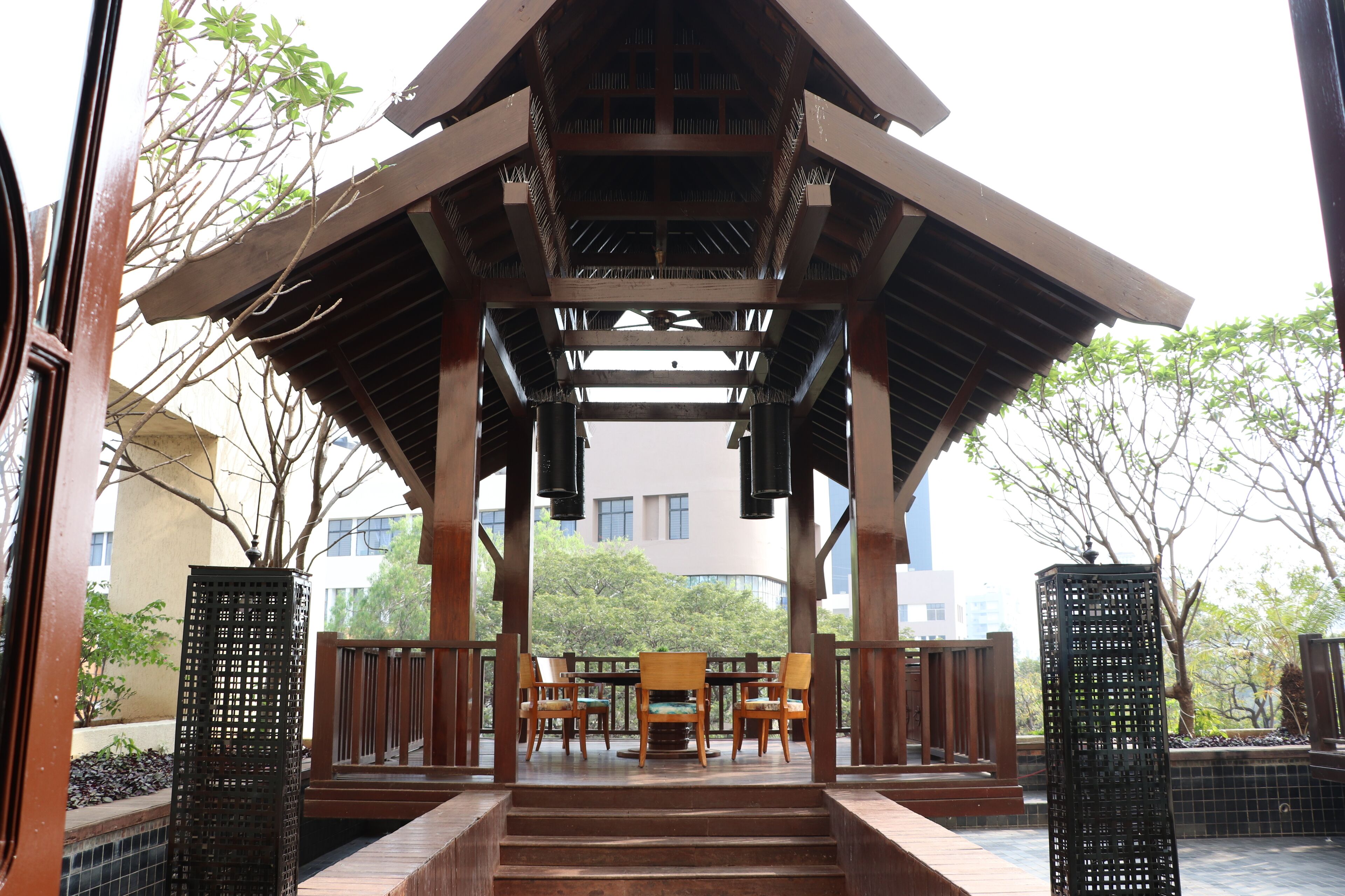 A covered outdoor seating area with wooden chairs and a table, surrounded by a wooden railing and a tiled floor.