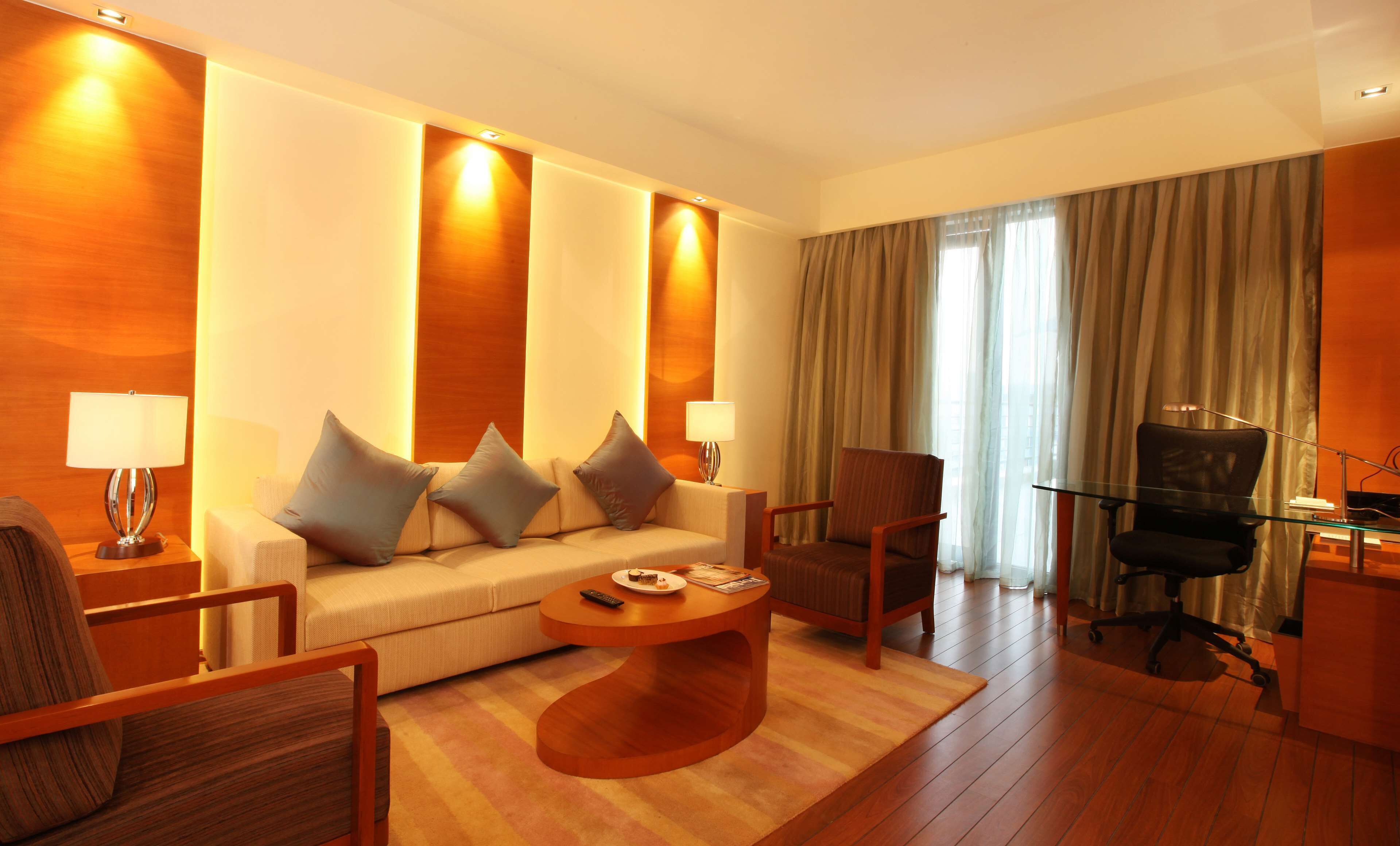 A modern hotel room with a sofa, armchairs, a coffee table, a desk with a chair, and a large window with curtains.