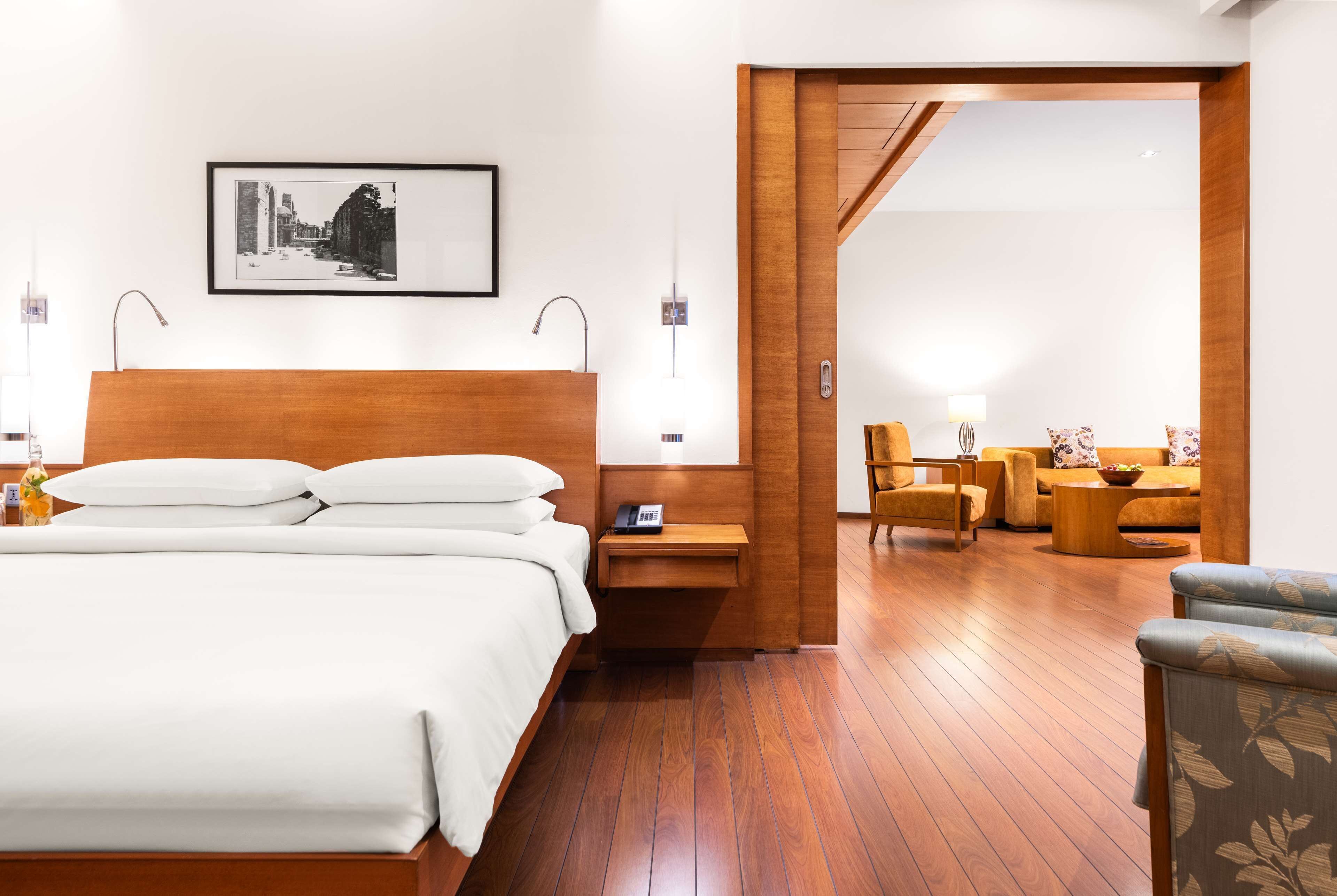 Une chambre d’hôtel avec un lit en bois, une table de chevet avec une lampe, un tableau encadré au mur et une vue sur un espace de vie comprenant un canapé et des fauteuils.