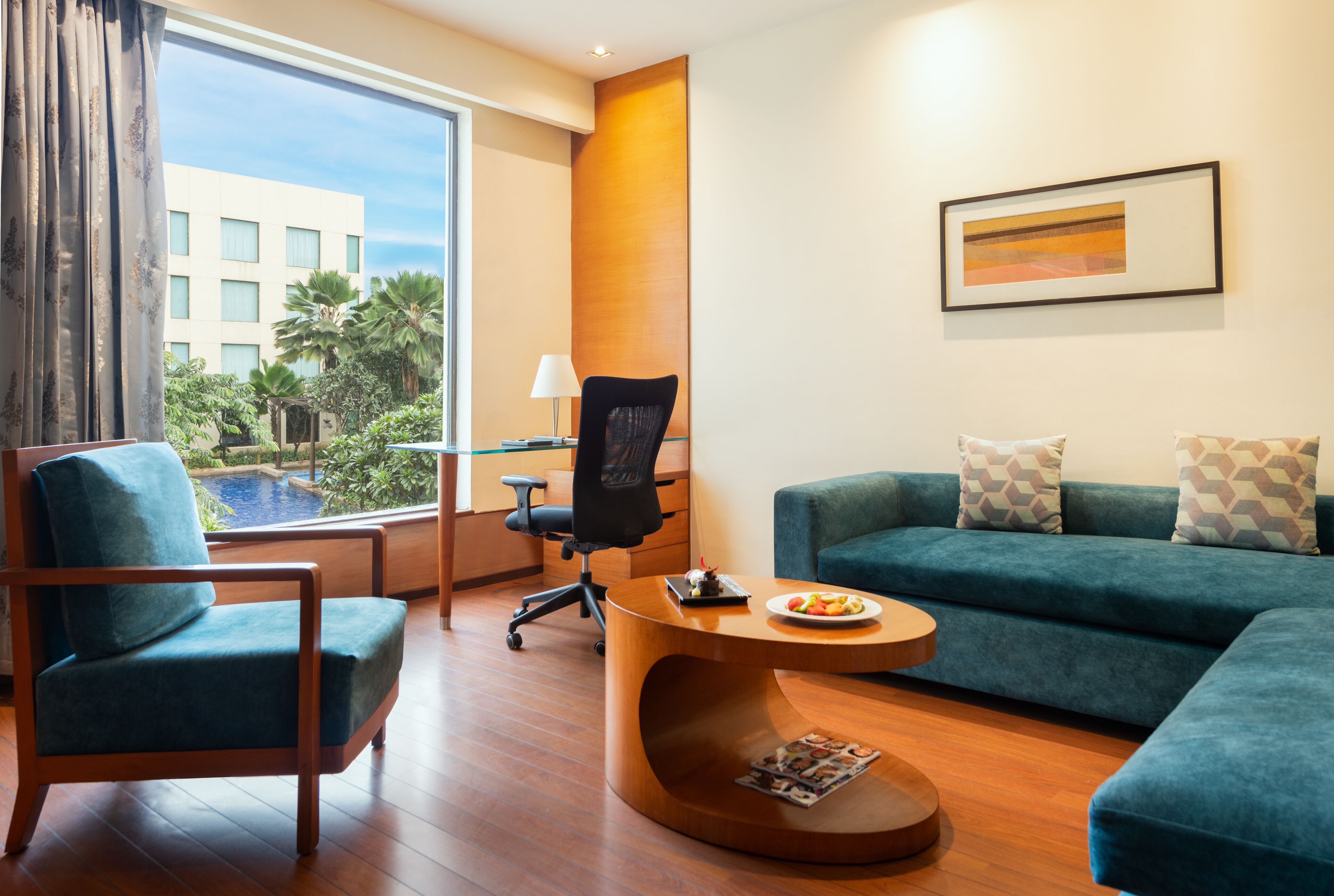 Quarto de hotel moderno com uma grande janela, um sofá azul, uma mesa de centro de madeira e uma cadeira de escritório preta.