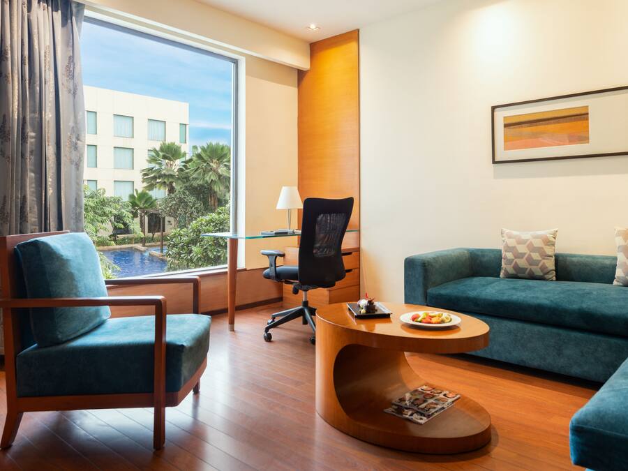 Una habitación de hotel moderna con un ventanal grande, un sofá azul, una mesa de centro de madera y una silla de oficina negra.