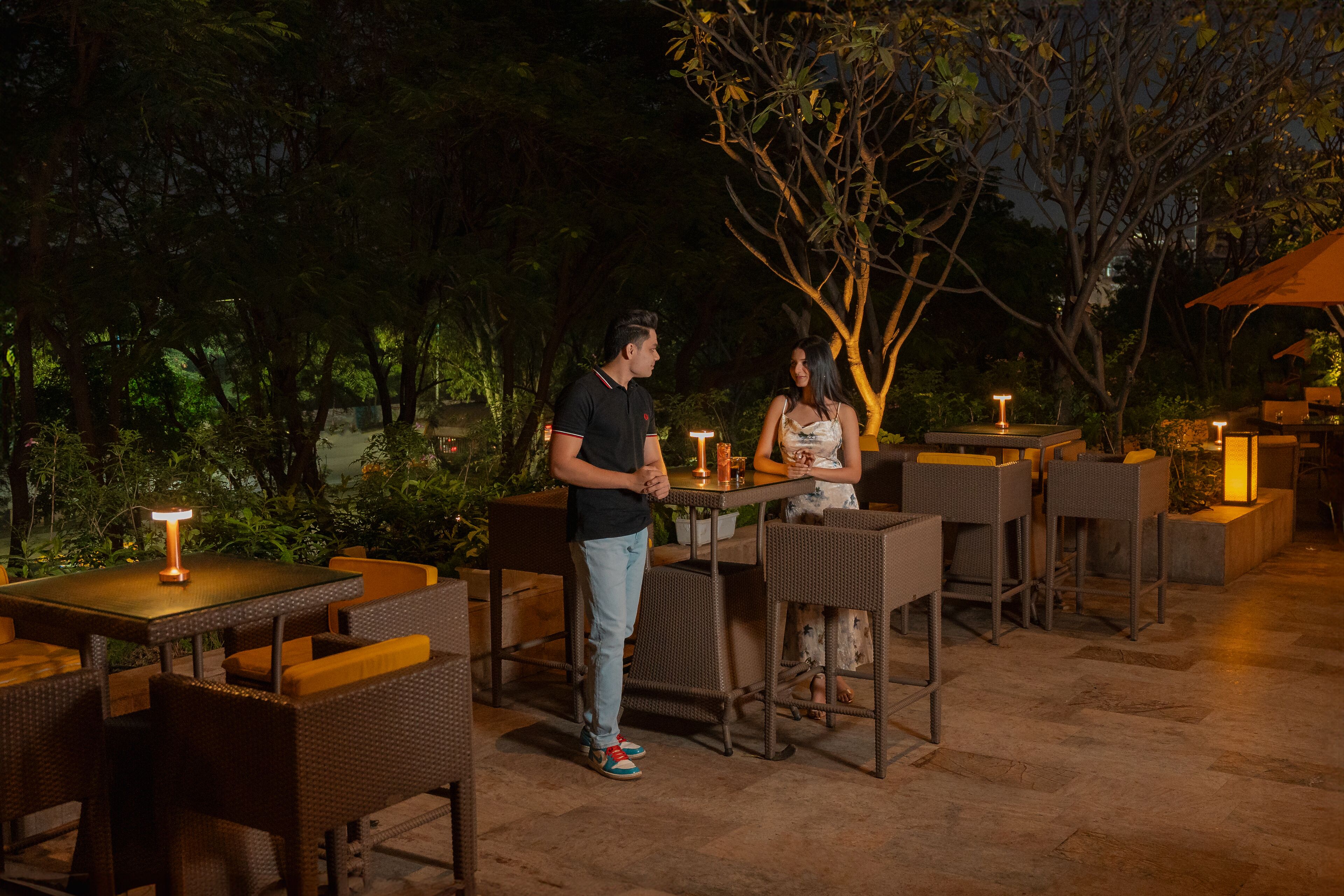 An outdoor dining area with tables and chairs, surrounded by trees and illuminated by table lamps.