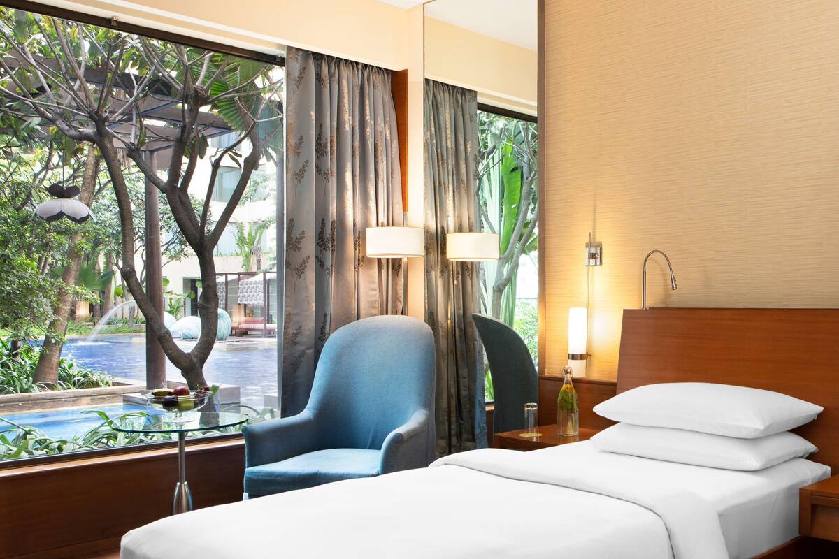 A hotel room with a bed, a chair, a glass table with fruits, and a view of an outdoor pool and greenery.