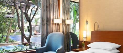 A hotel room with a bed, a chair, a glass table with fruits, and a view of an outdoor pool and greenery.