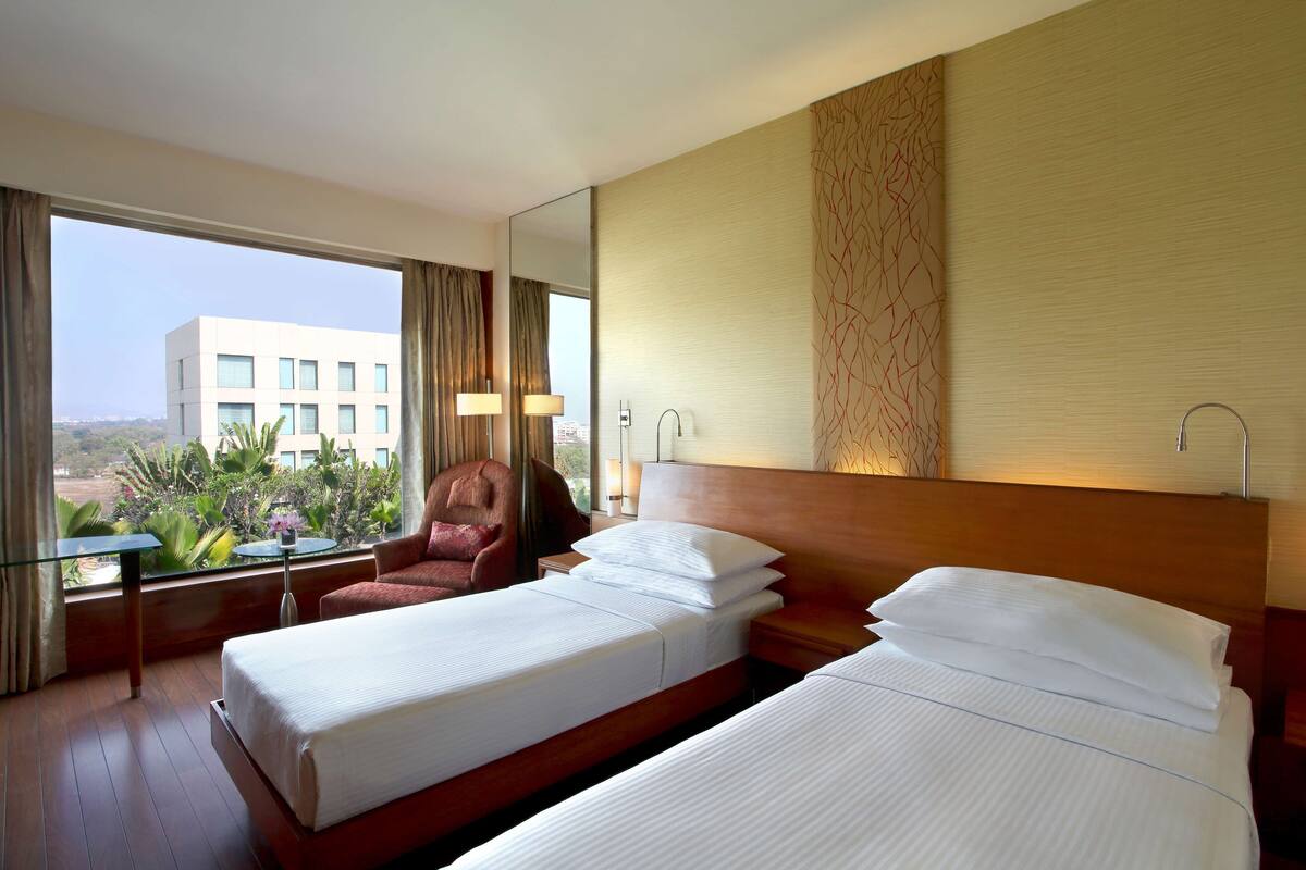 A hotel room with two beds, a chair, a table, and a view of a building and greenery outside.