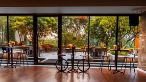An indoor seating area with wooden flooring, tables, and chairs, large windows with a view of greenery and trees. - Hyatt Pune (Pune)
