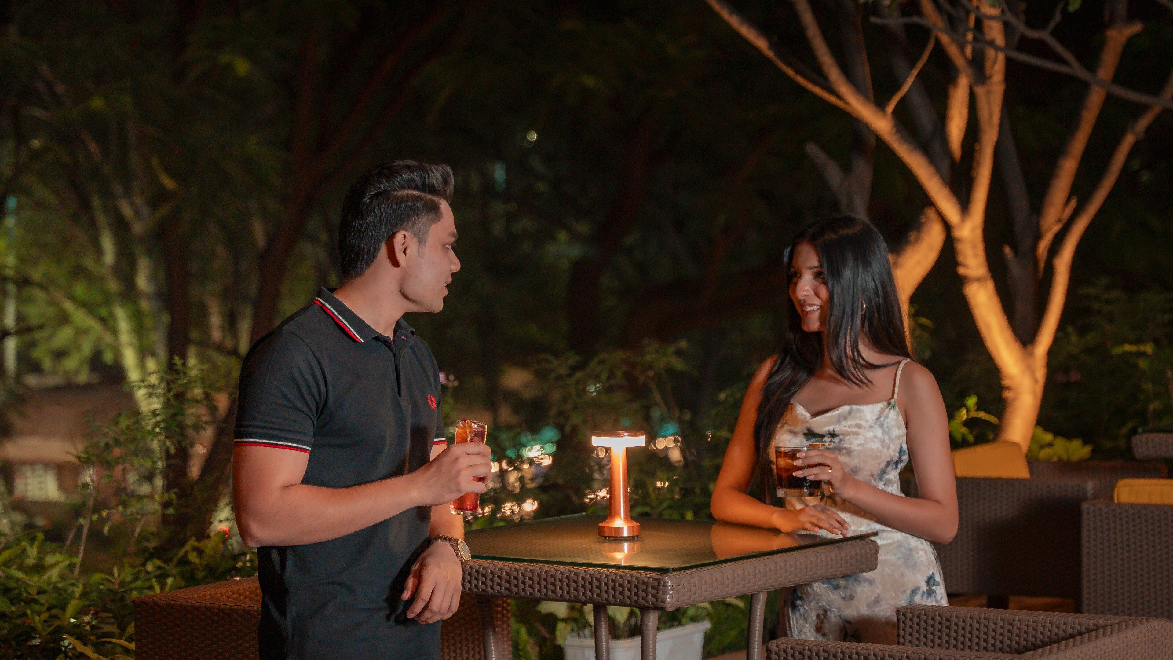 Two people standing at an outdoor table with a candle in the center, surrounded by greenery.
