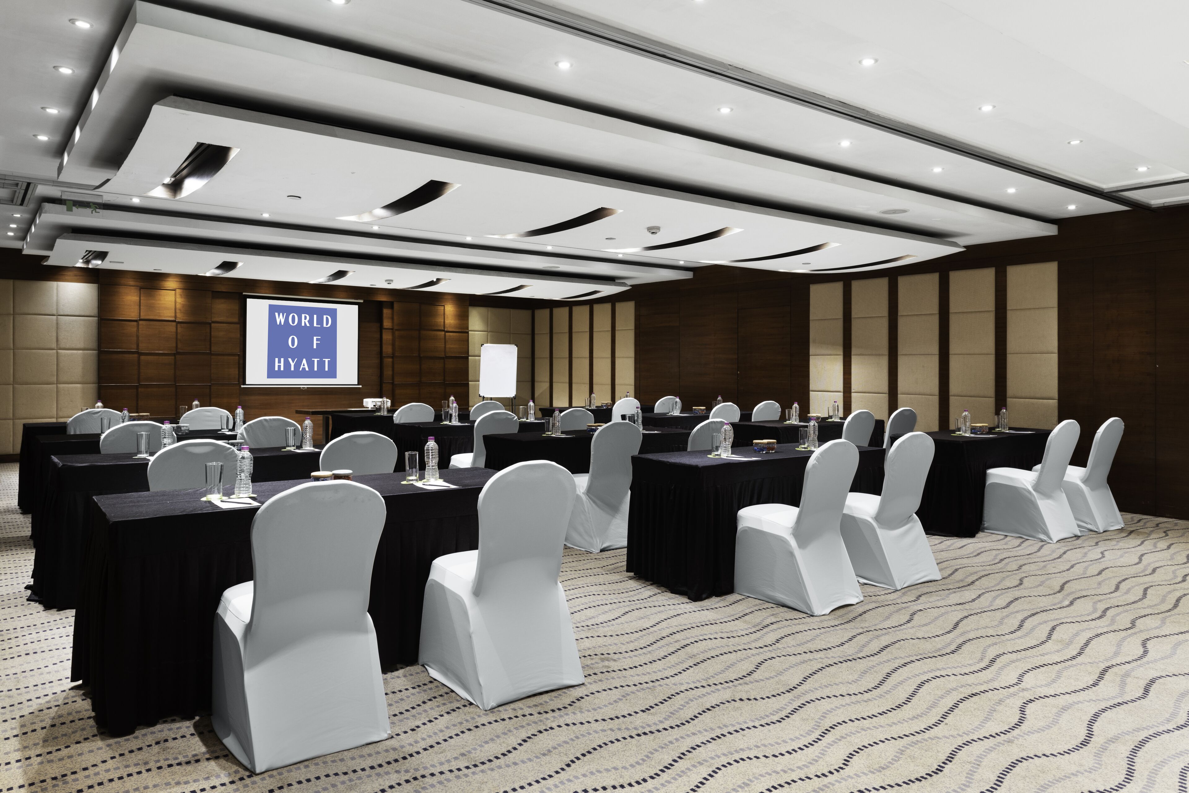 Un salón de conferencias con varias mesas, sillas blancas y una pantalla grande que muestra “WORLD OF HYATT”.