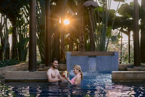 Una pareja en una piscina con fondo de azulejos y plantas tropicales.