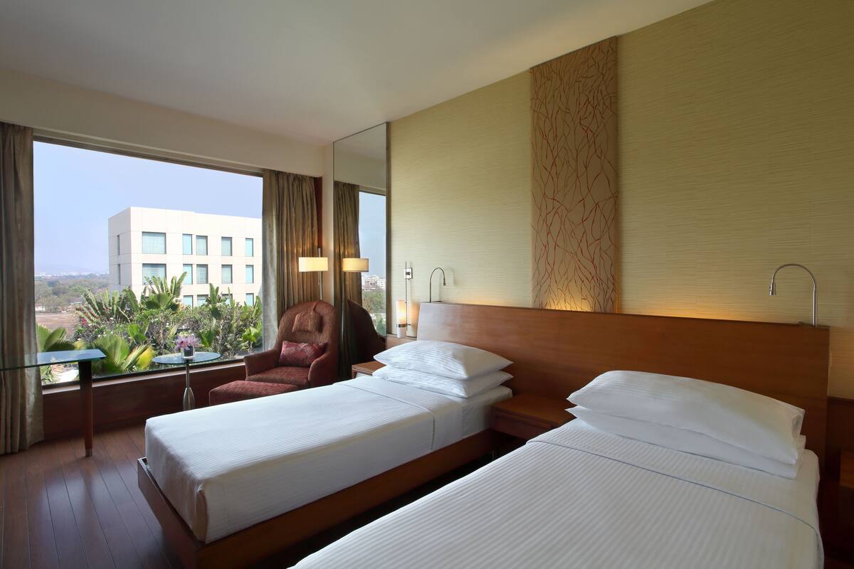 A hotel room with two beds, a large window with a view of a building and greenery, a chair, and a table.
