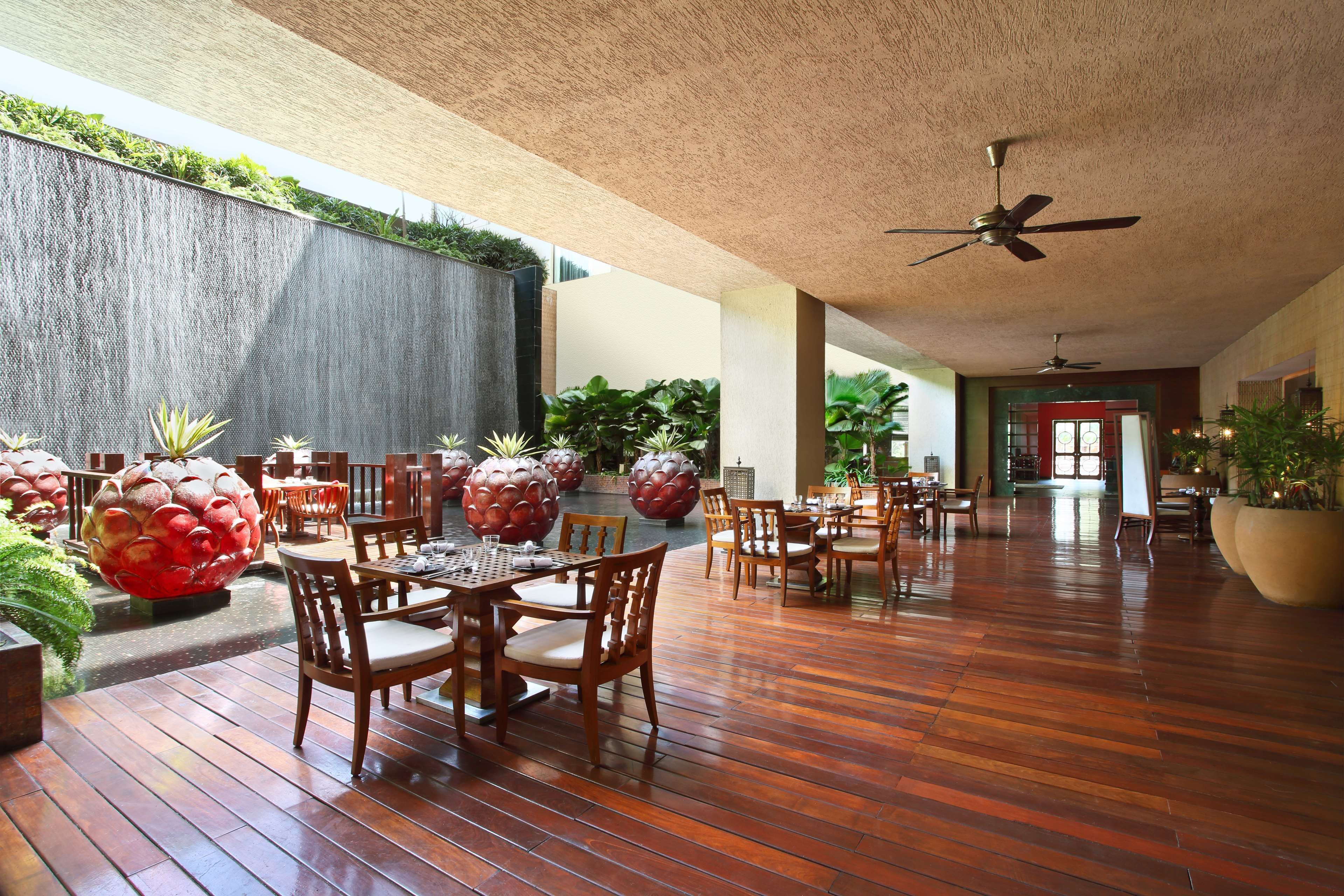 Um espaço amplo e interno com piso de madeira, várias mesas e cadeiras para refeições, ventiladores de teto e plantas em vasos.