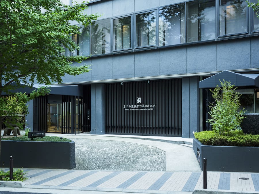 A modern hotel entrance with a dark facade, large windows, and a sign in a foreign language.