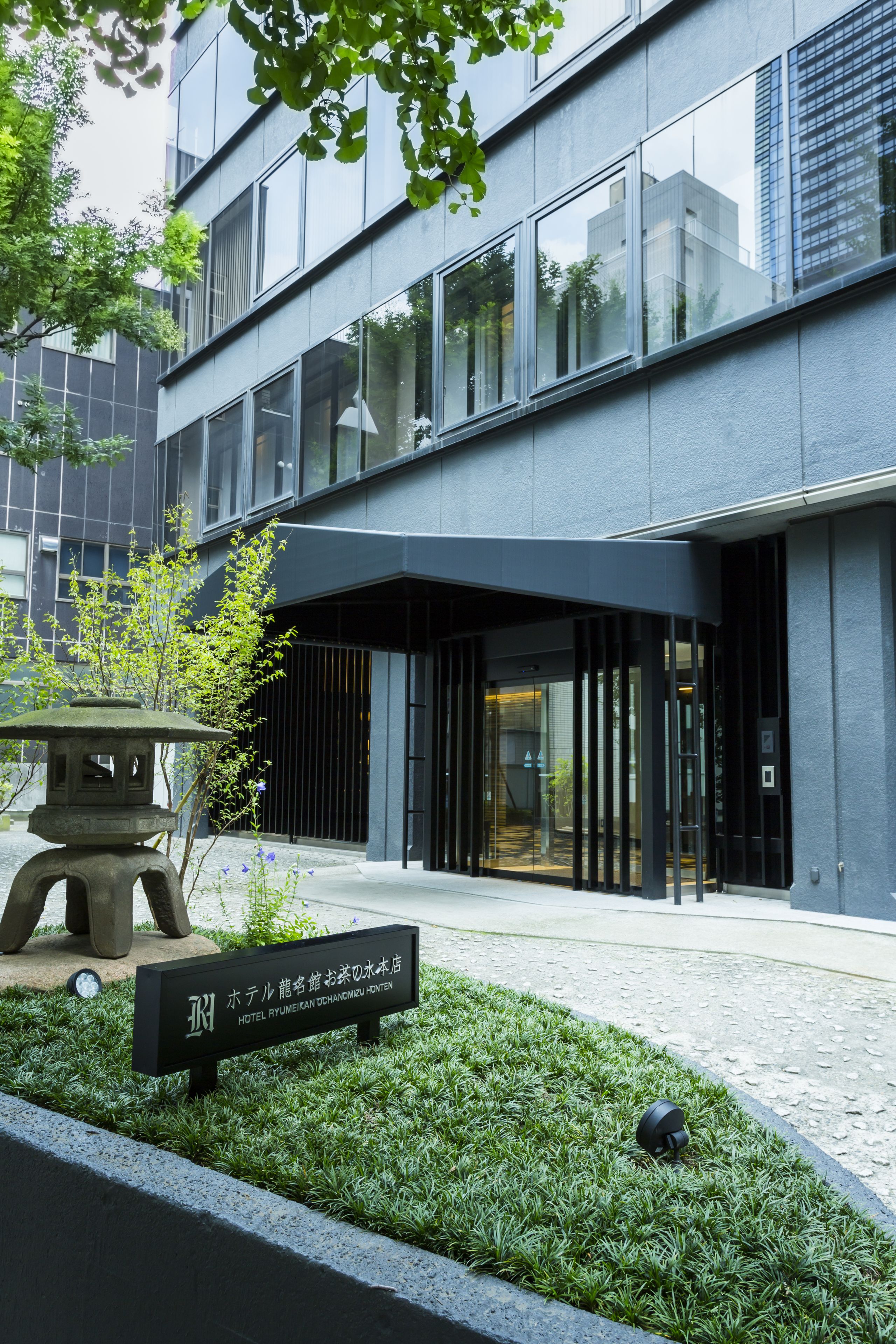 A modern hotel entrance with a clear glass facade, a stone lantern, and a neatly trimmed lawn.