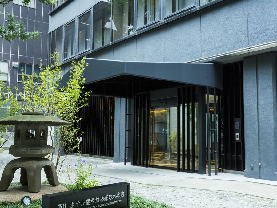 A modern hotel entrance with a clear glass facade, a stone lantern, and a neatly trimmed lawn.