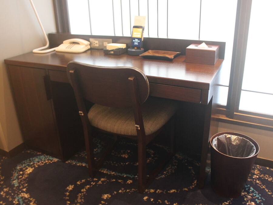 A hotel room with a wooden desk, a telephone, a mobile phone charging station, a tissue box, and a trash can.