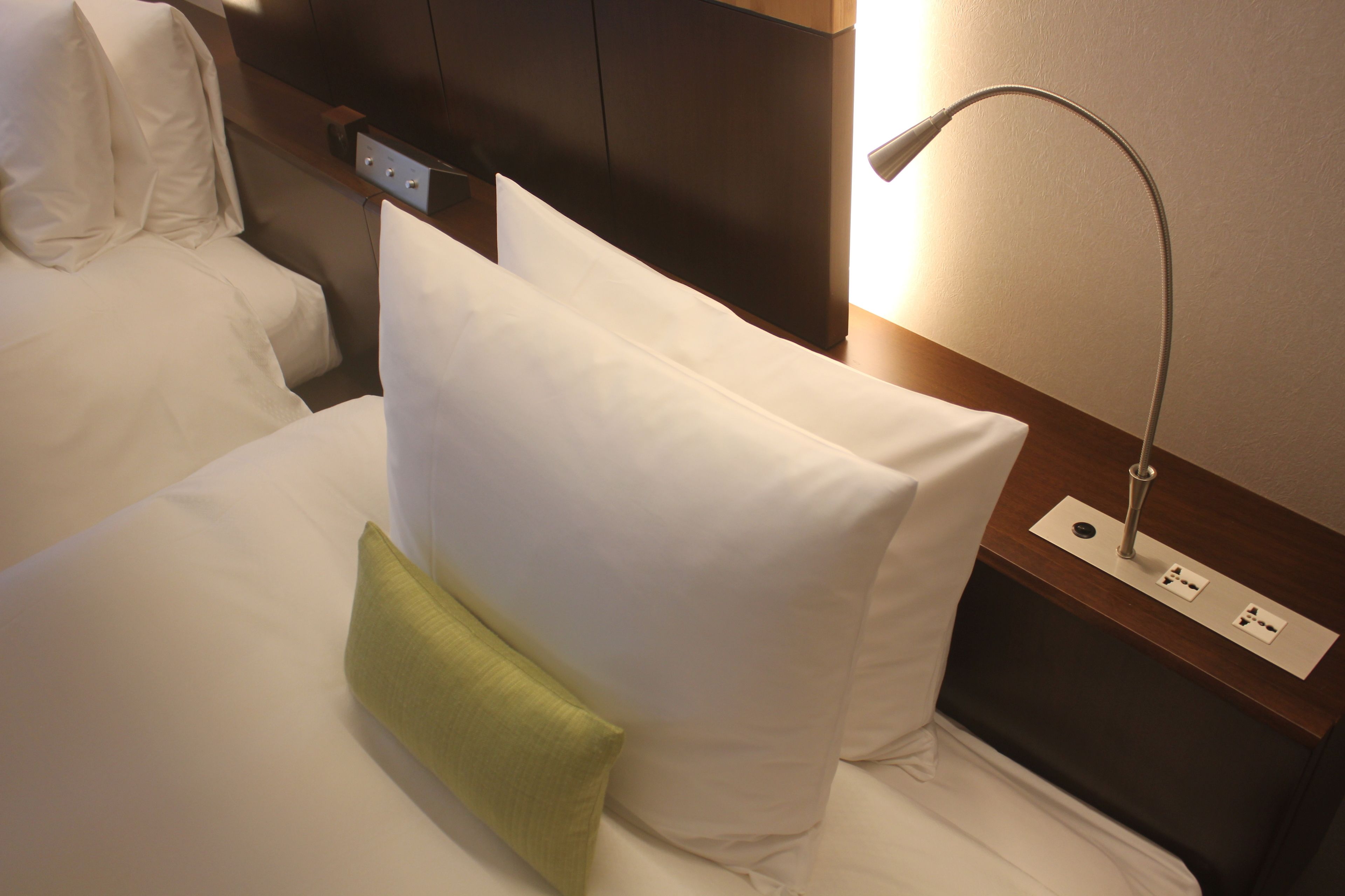 Habitación de hotel con una cama bien hecha, una mesita de noche con lámpara y un enchufe en la pared.