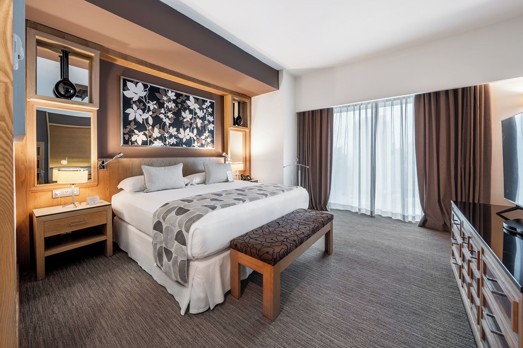 Habitación de hotel con una cama grande, una mesita de noche con lámpara, un banco, un televisor y un ventanal con cortinas.