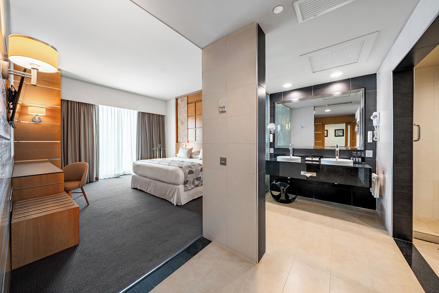 A modern hotel room with a bed, bedside table, lamp, and a view of the hallway.