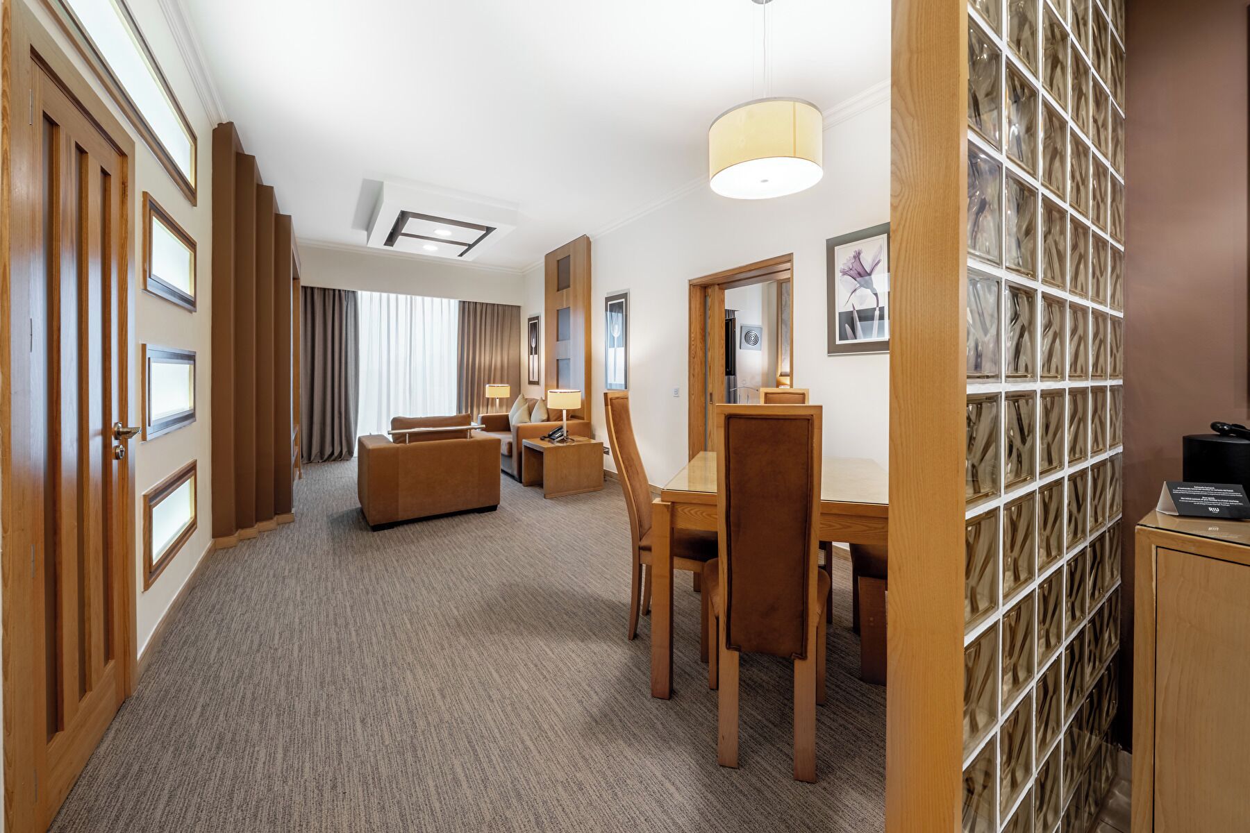 A hotel room with a seating area, a desk, a lamp, and a bookshelf.