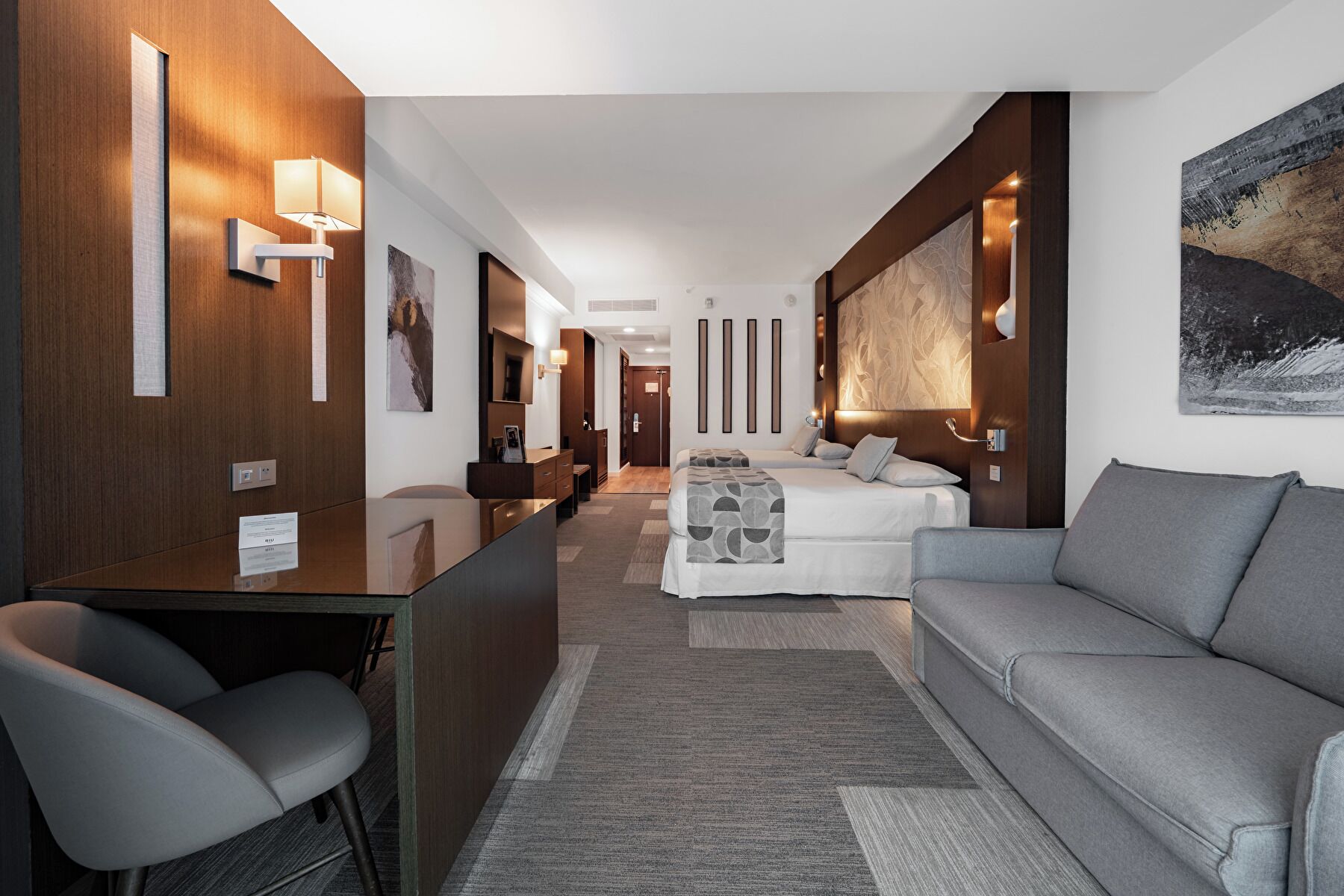 A modern hotel room with a sofa, a desk, and a bed. There are wall-mounted lights and a painting on the wall.
