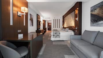 A modern hotel room with a sofa, a desk, and a bed. There are wall-mounted lights and a painting on the wall.