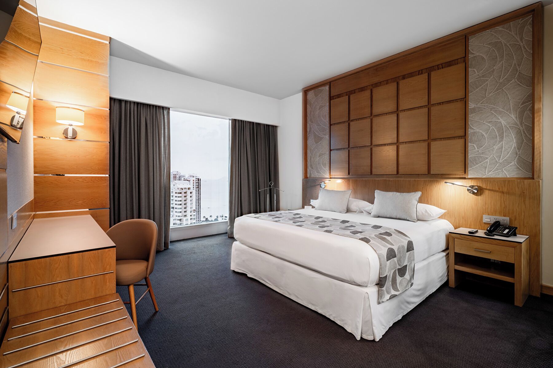 A hotel room with a large bed, a desk, a chair, a nightstand, and a view of the city through the window.