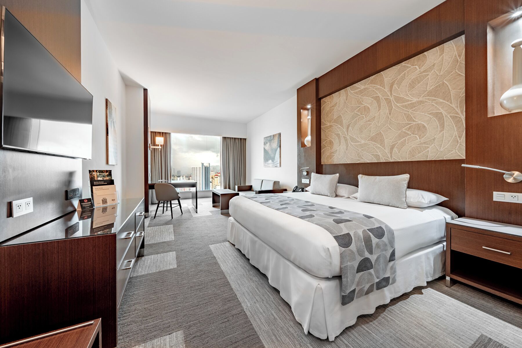 A modern hotel room with a large bed, a desk, a chair, and a TV.