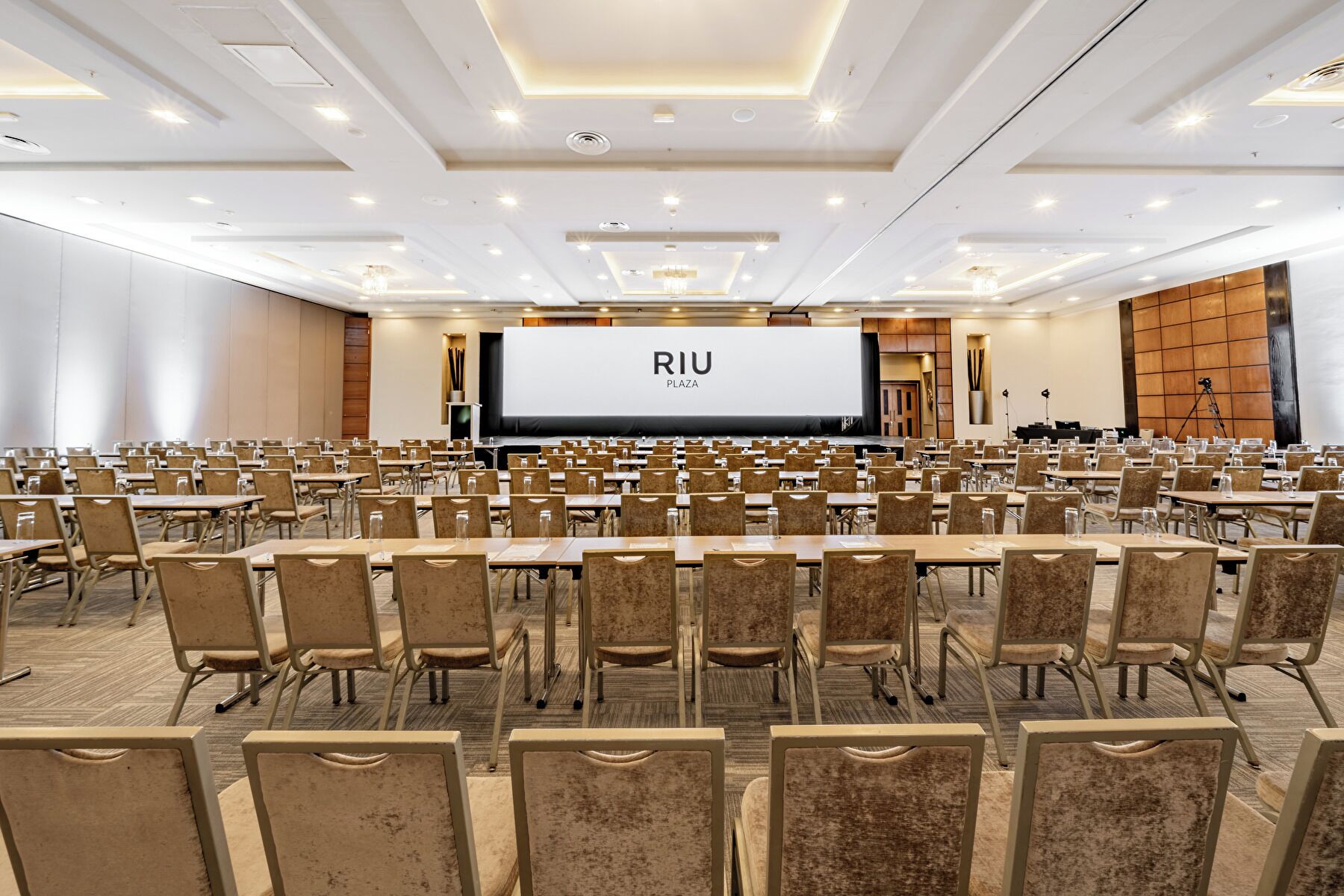 A conference room with rows of tables and chairs, a large screen displaying "RIU PLAZA", and a stage with a podium.