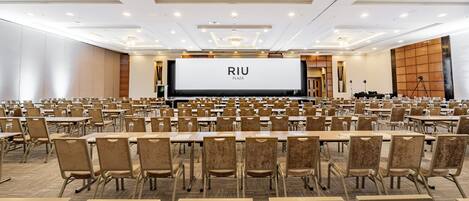 A conference room with rows of tables and chairs, a large screen displaying "RIU PLAZA", and a stage with a podium.