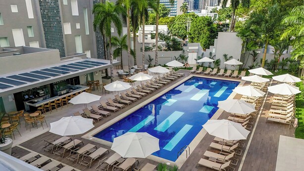 A rooftop pool with sun loungers and umbrellas, surrounded by palm trees and modern buildings.
