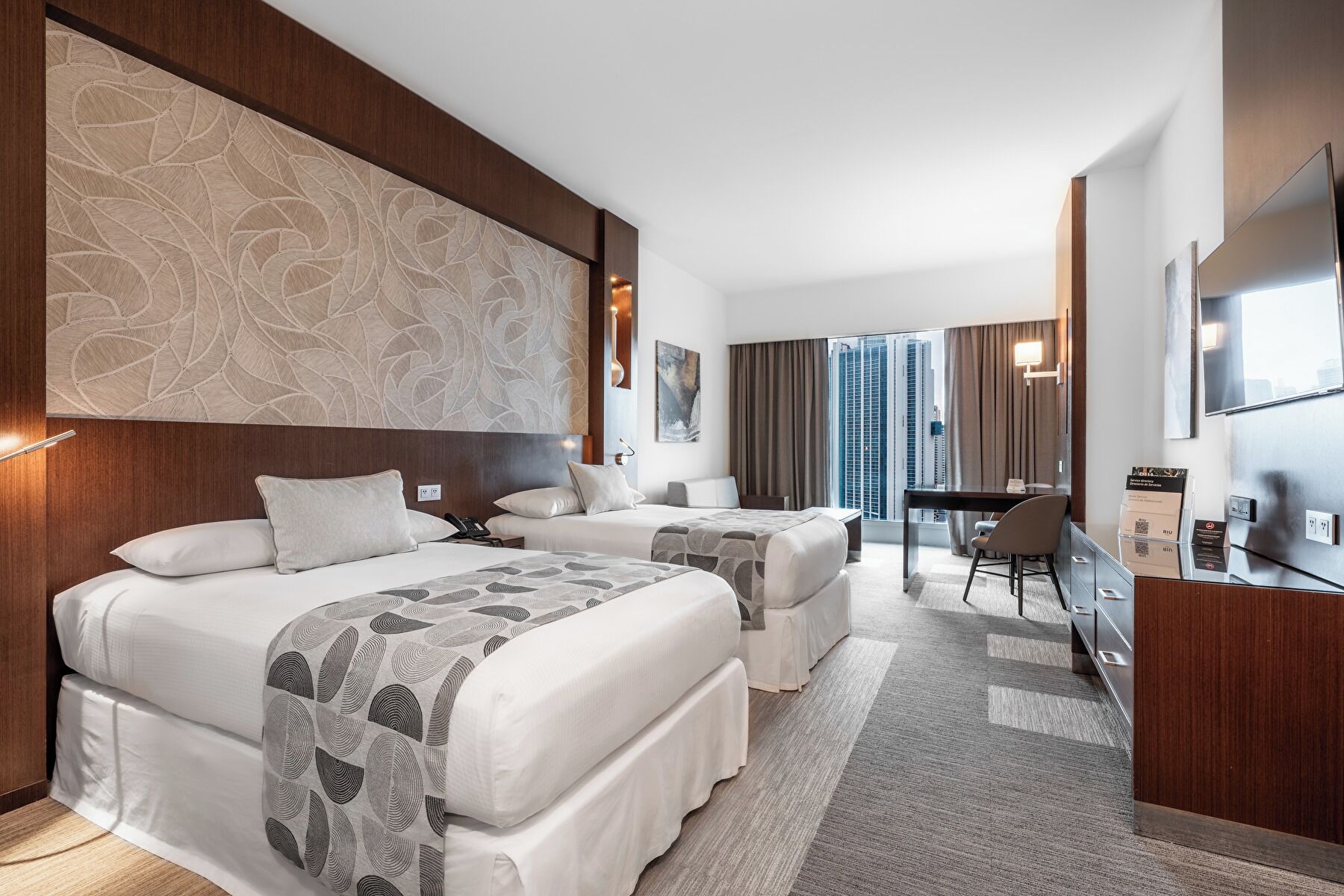 A hotel room with two beds, a desk, a chair, and a large window offering a city view.