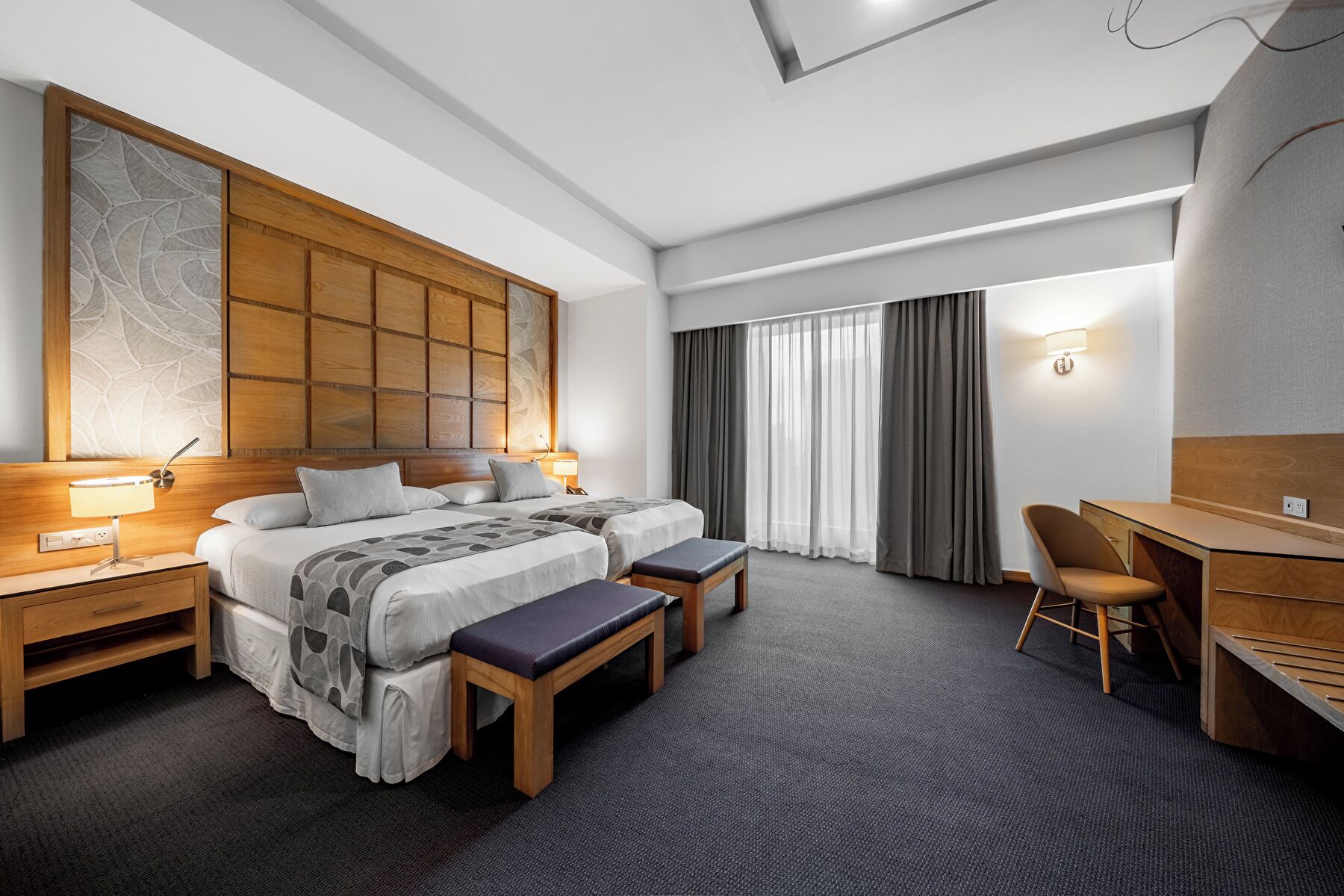 A hotel room with a large bed, two bedside tables, a desk, a chair, and a large window with curtains.